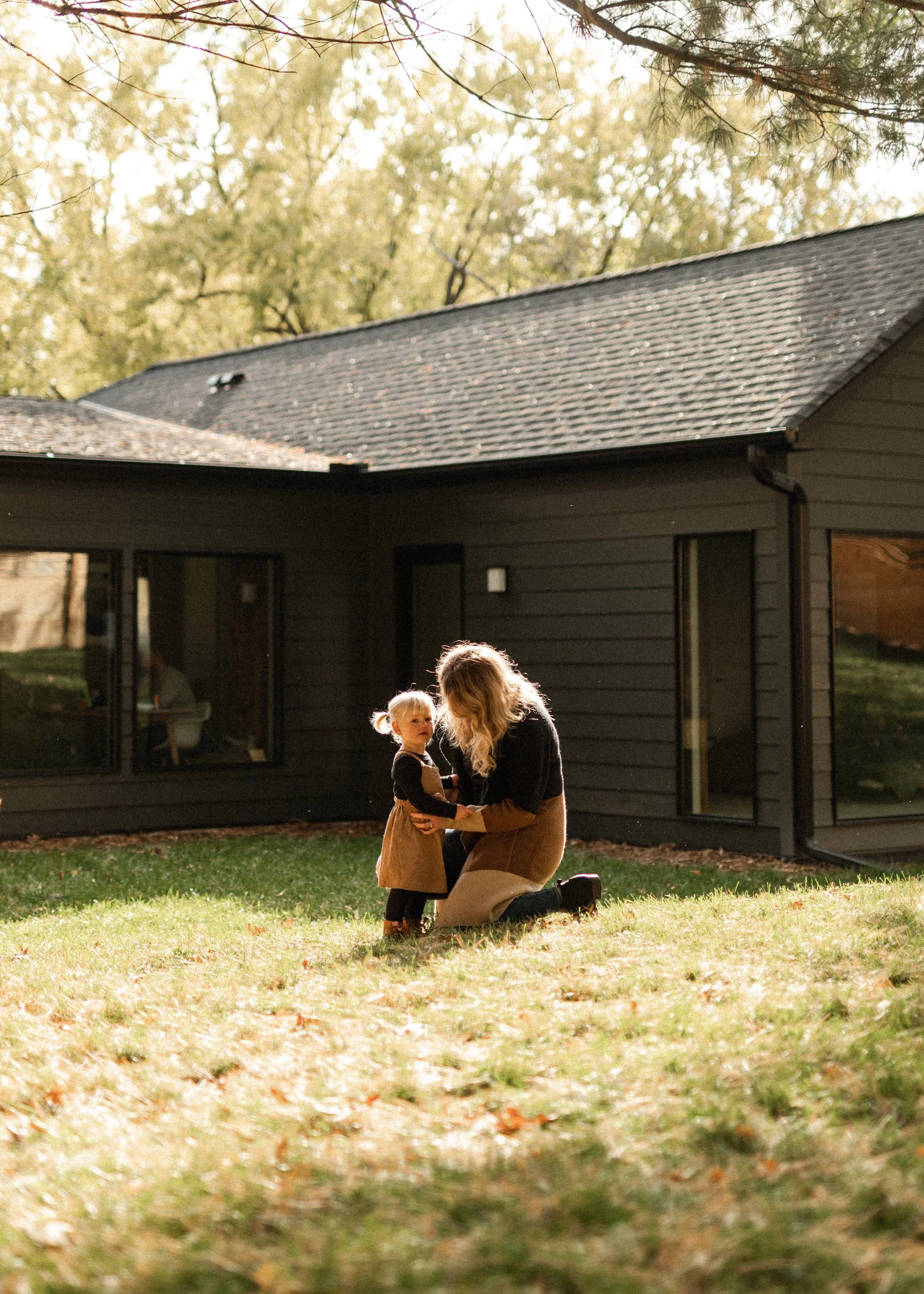 A woman and a young girl are kneeling on the grass outside a dark-colored house, facing each other and holding hands. The woman has long blonde hair, and the girl has blonde hair styled in a ponytail. The background features a house with large window