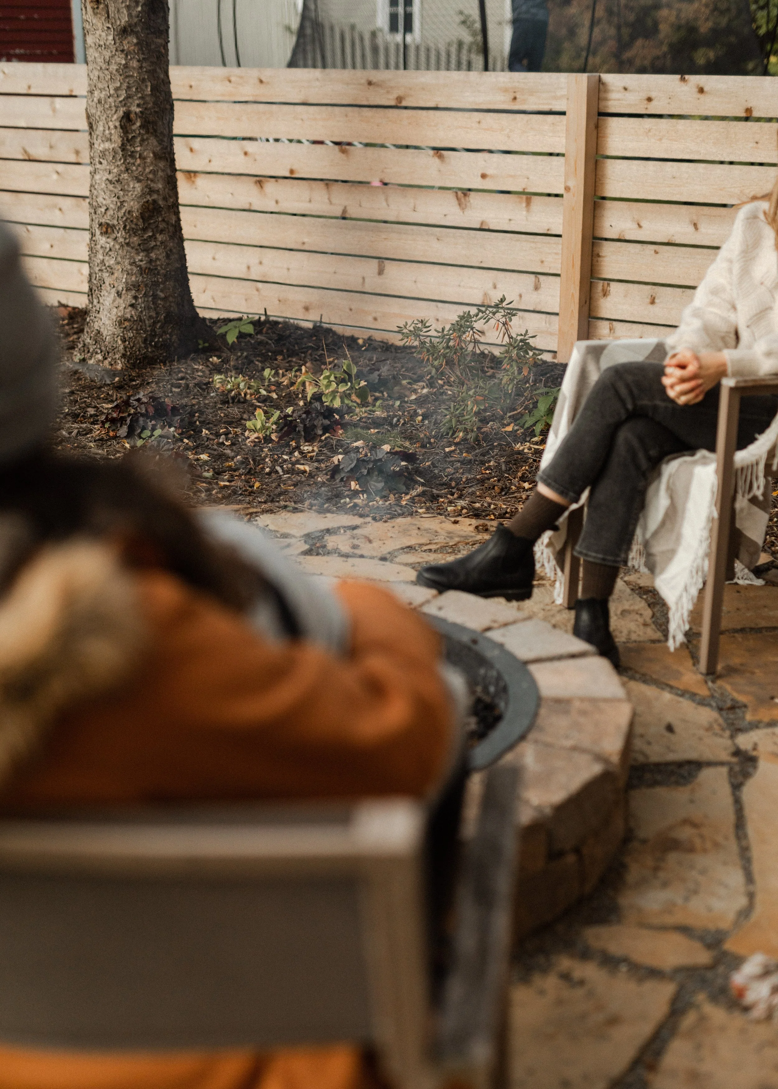 A wooden privacy fence under construction with a tree trunk and some small plants in the garden area beside it, and a person wearing white sweater and black boots sitting on a chair.