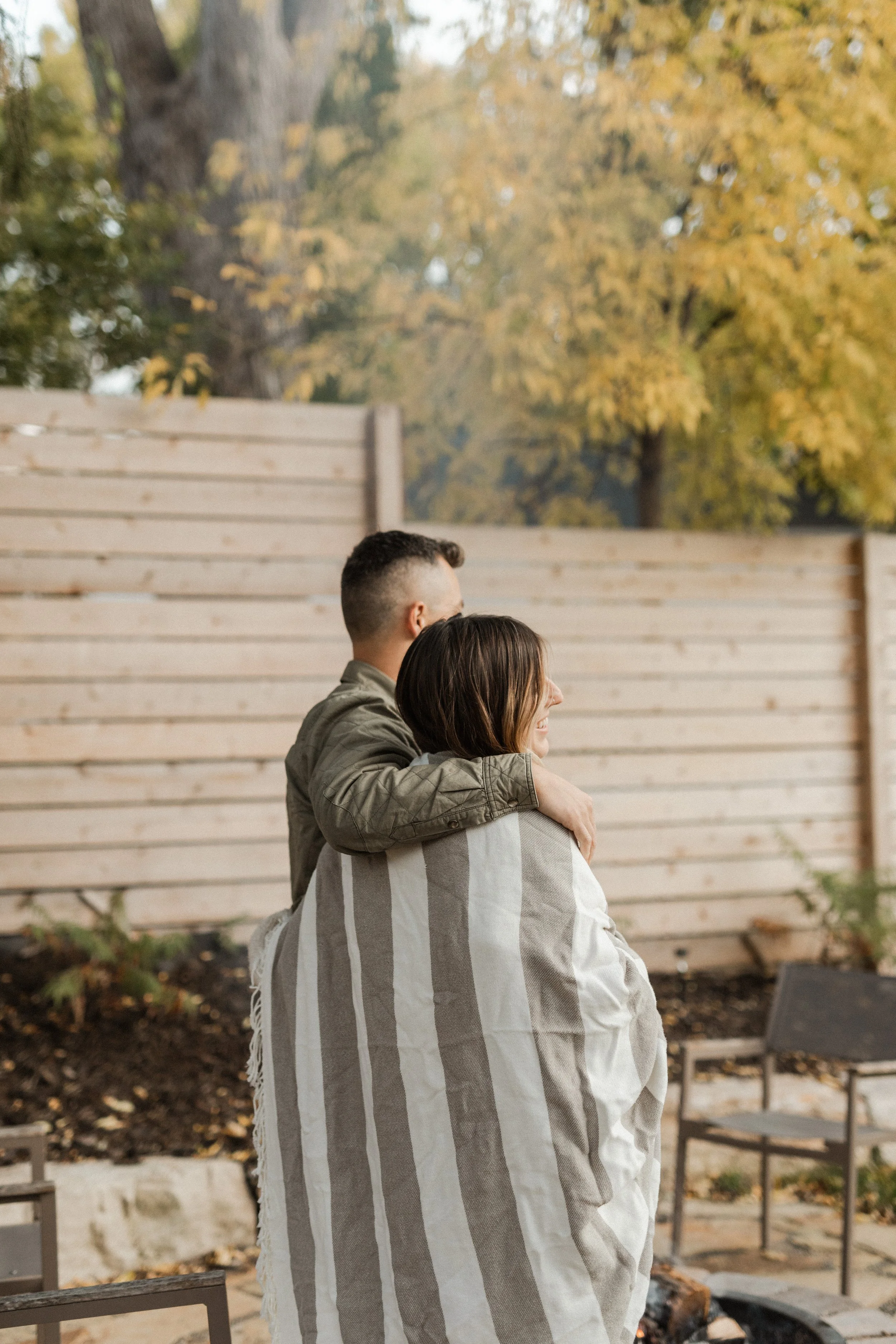 A couple standing outdoors in front of a wooden fence, with trees showing autumn colors in the background. The man is hugging the woman.