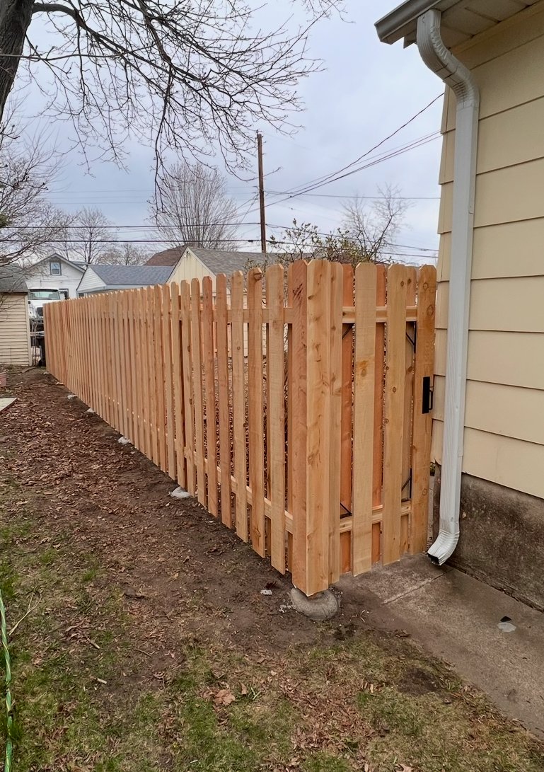 View of a new wooden fence installed along the side of a house with beige siding, showing a small gate, dirt yard, and overcast sky.