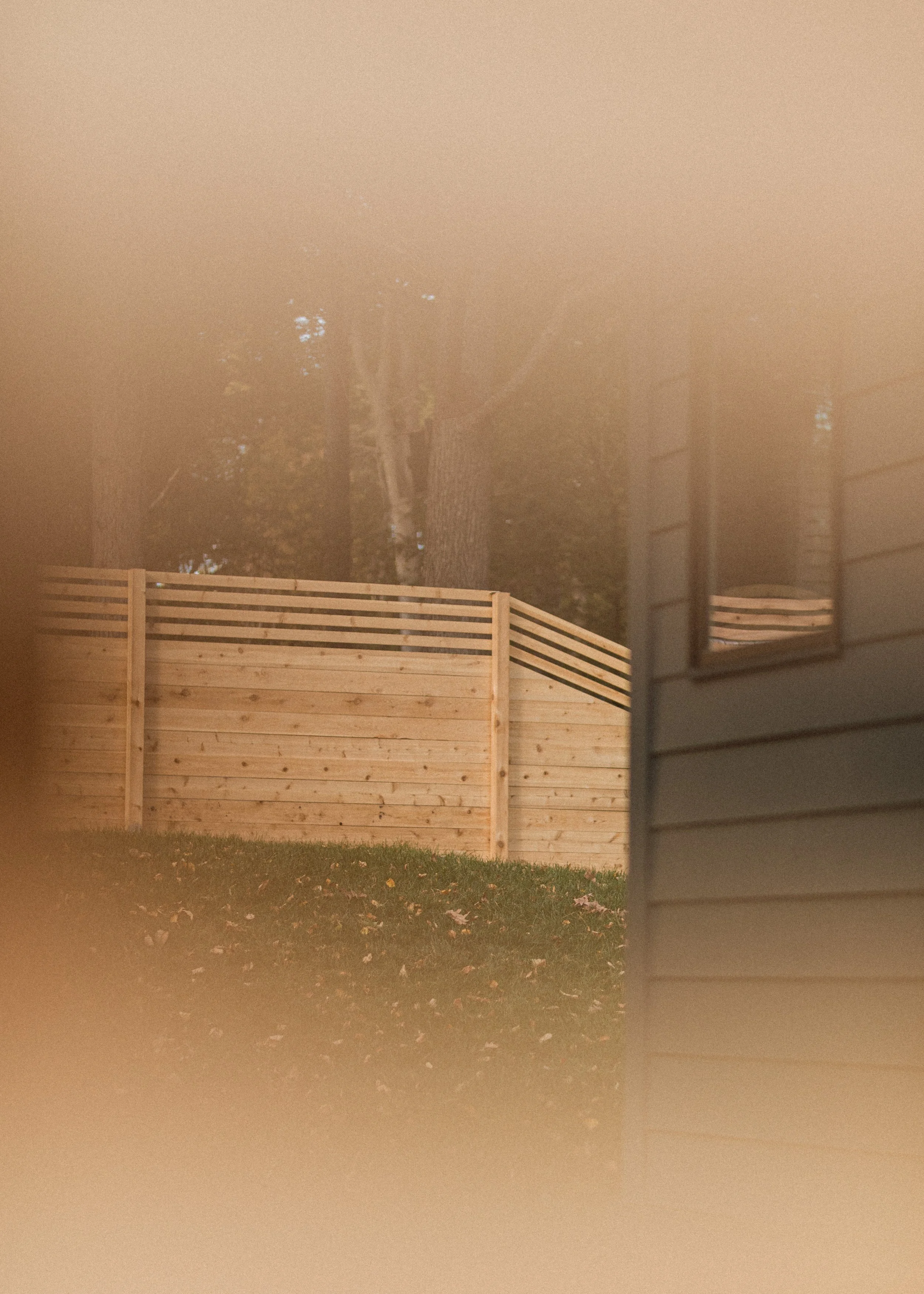 A newly built wood fence with horizontal slats stands in a yard. Behind the fence, there's a large tree with a tall trunk and branches. Part of a house or building with siding is visible to the right, and the scene is viewed through a window with a p