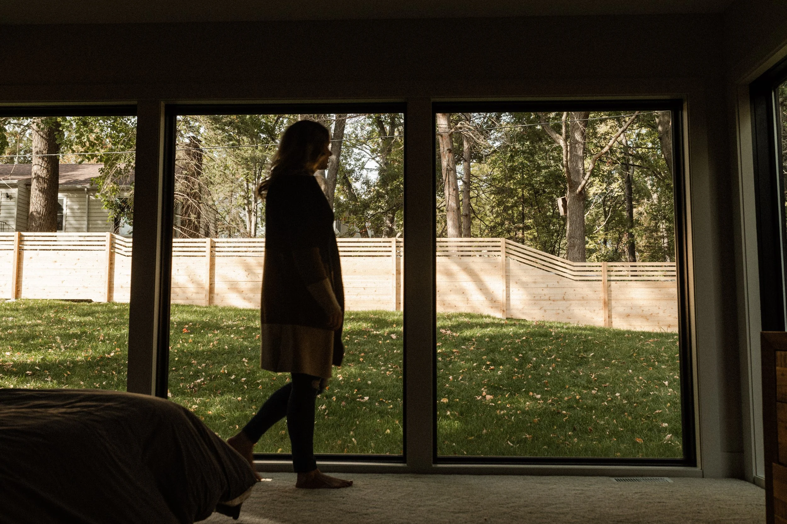 A person walking outside in a backyard with green grass and trees, viewed from inside a house through sliding glass doors.