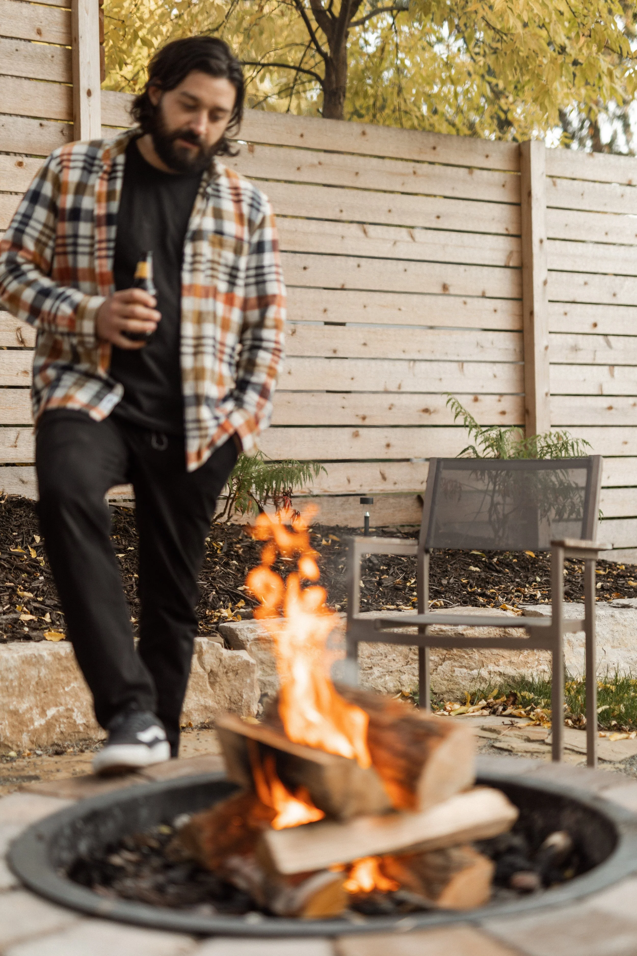 A man with long hair and a beard wearing a checkered flannel shirt stands outdoors by a fire pit, holding a beer bottle, with a wooden fence and autumn trees in the background.