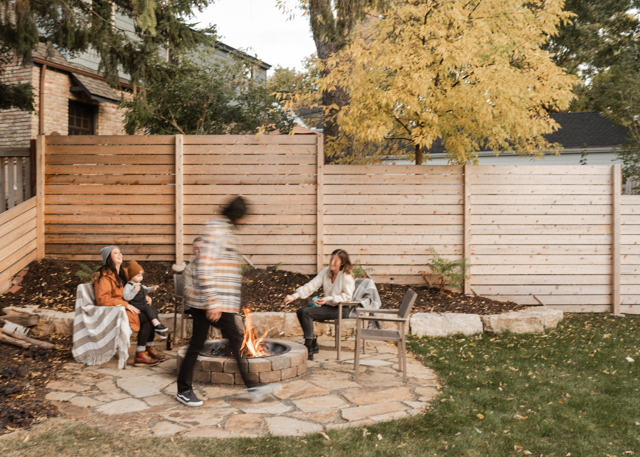 A family sitting around a fire pit in a backyard with autumn leaves on the ground and a wooden fence. A large tree with yellow leaves is visible in the background. Several people are gathered, with some standing and some sitting.