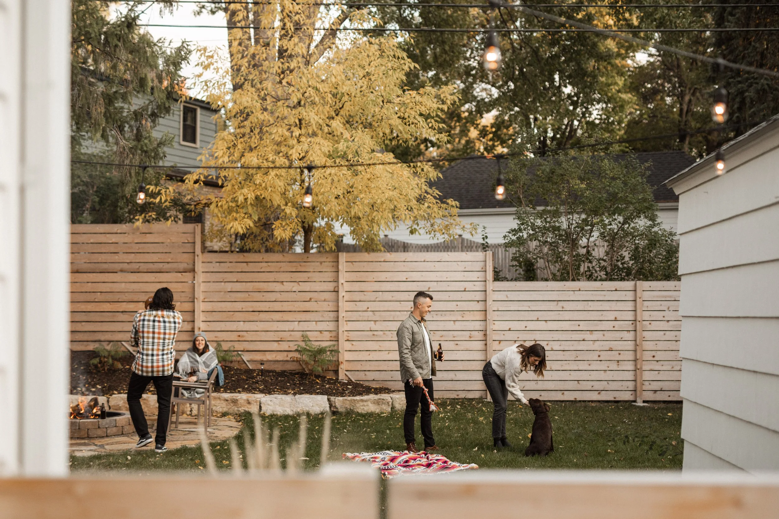 People socializing in a backyard during fall, with a wooden fence, trees with yellow leaves, string lights, and a fire pit.