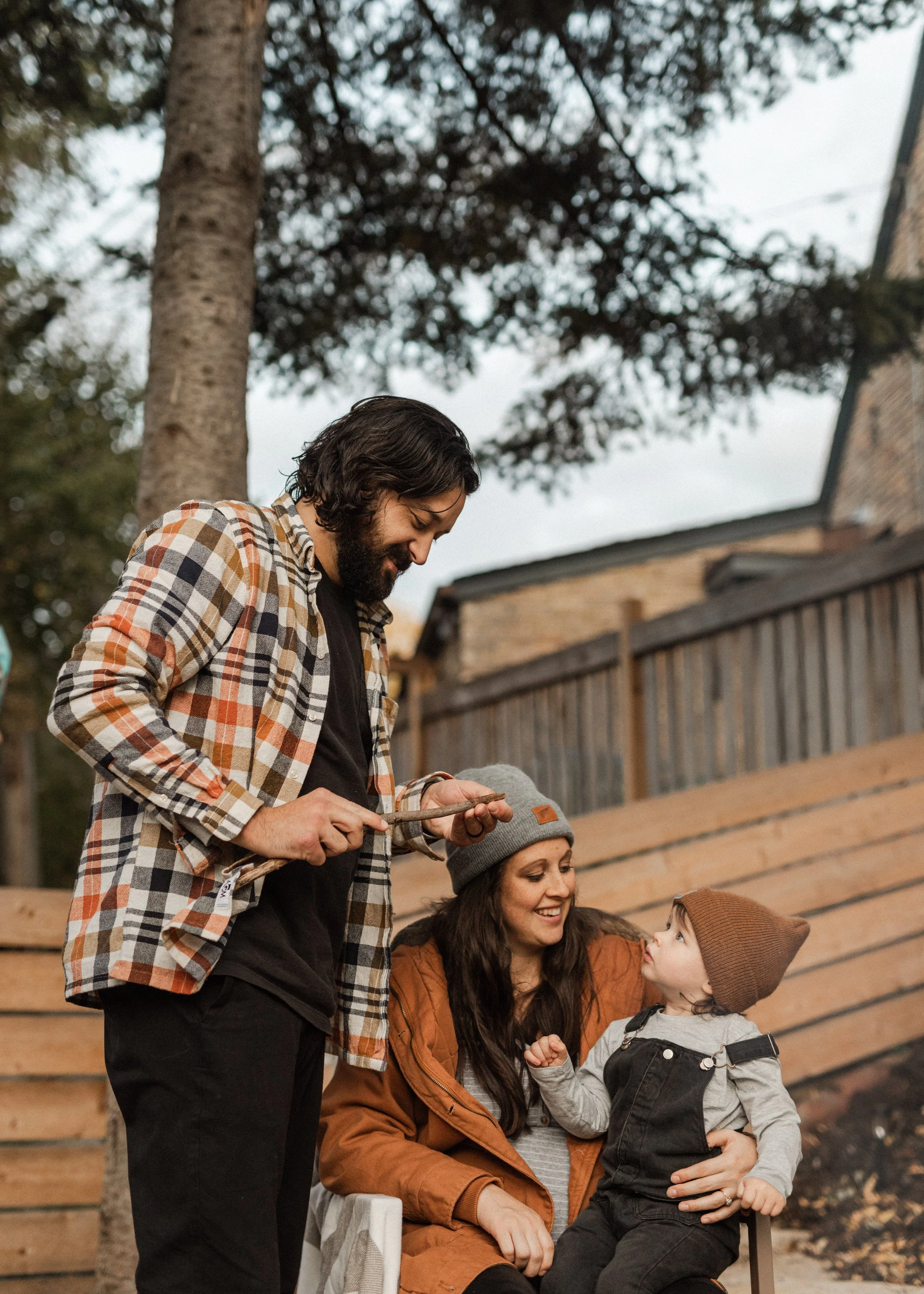 A group of people, including a man with dark hair and a beard, a woman with long dark hair, and a young boy, are outside near a wooden fence and tall trees. The man is standing, smiling, and looking down at the boy, who is sitting on a chair and wear