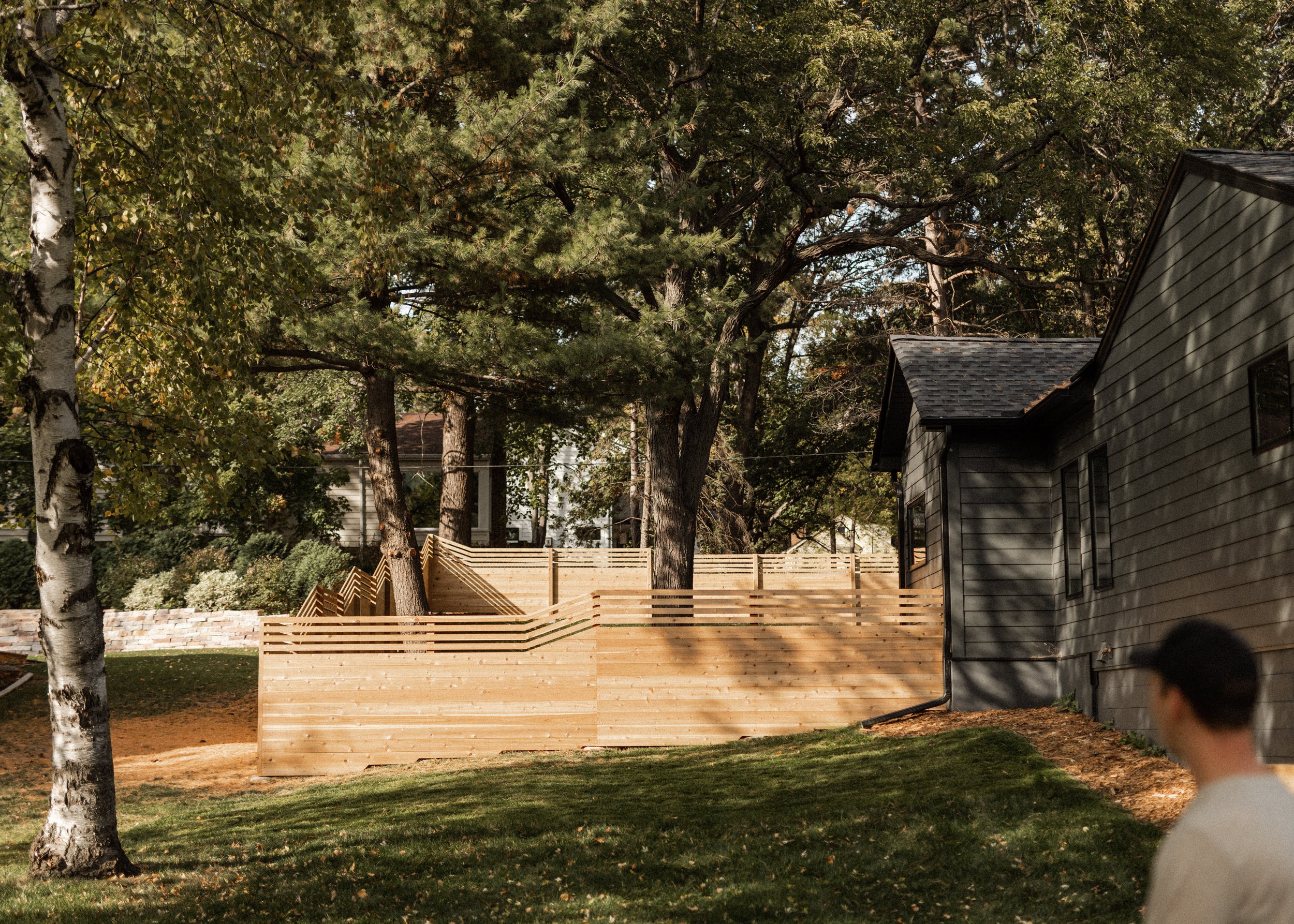 A backyard with tall trees, a gray house, a wooden privacy fence, and a person partially visible in the foreground.