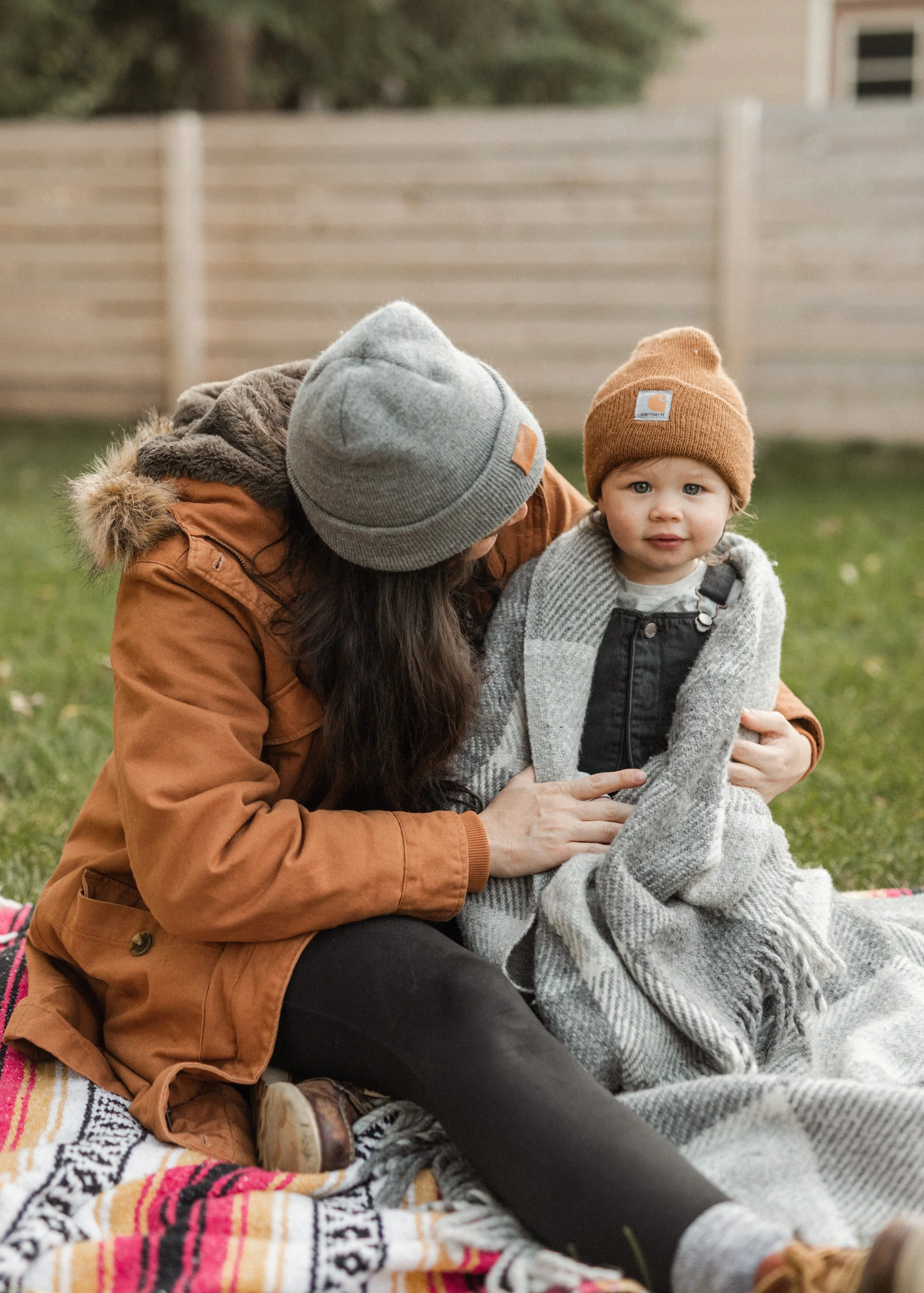 A woman and a young child sitting on a colorful blanket outdoors, wearing warm clothing and knit hats, with a wooden fence and trees in the background.