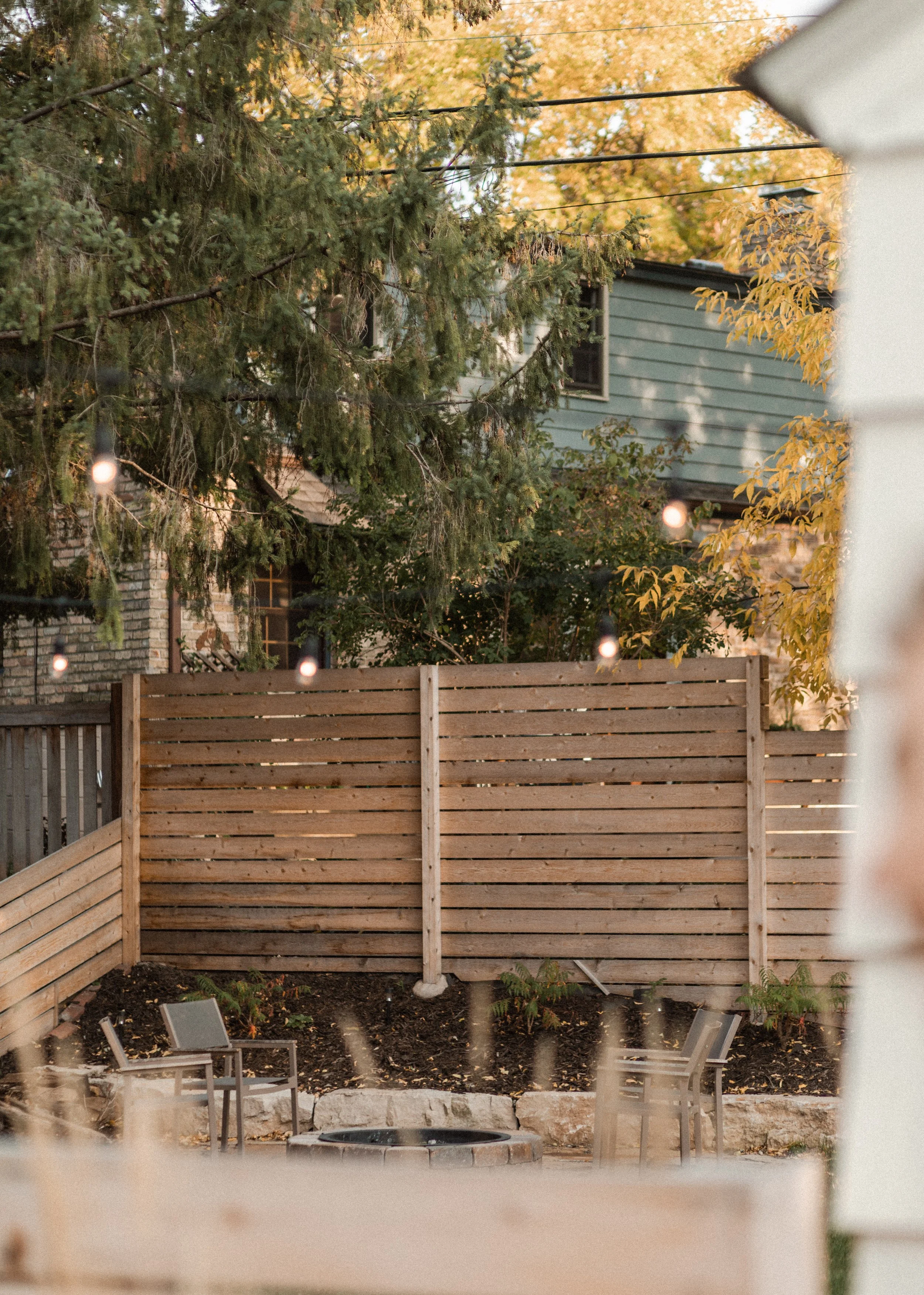 View of a backyard with a wooden privacy fence, a small fire pit, garden chairs, solar lights, trees with autumn leaves, and a house with gray siding in the background.