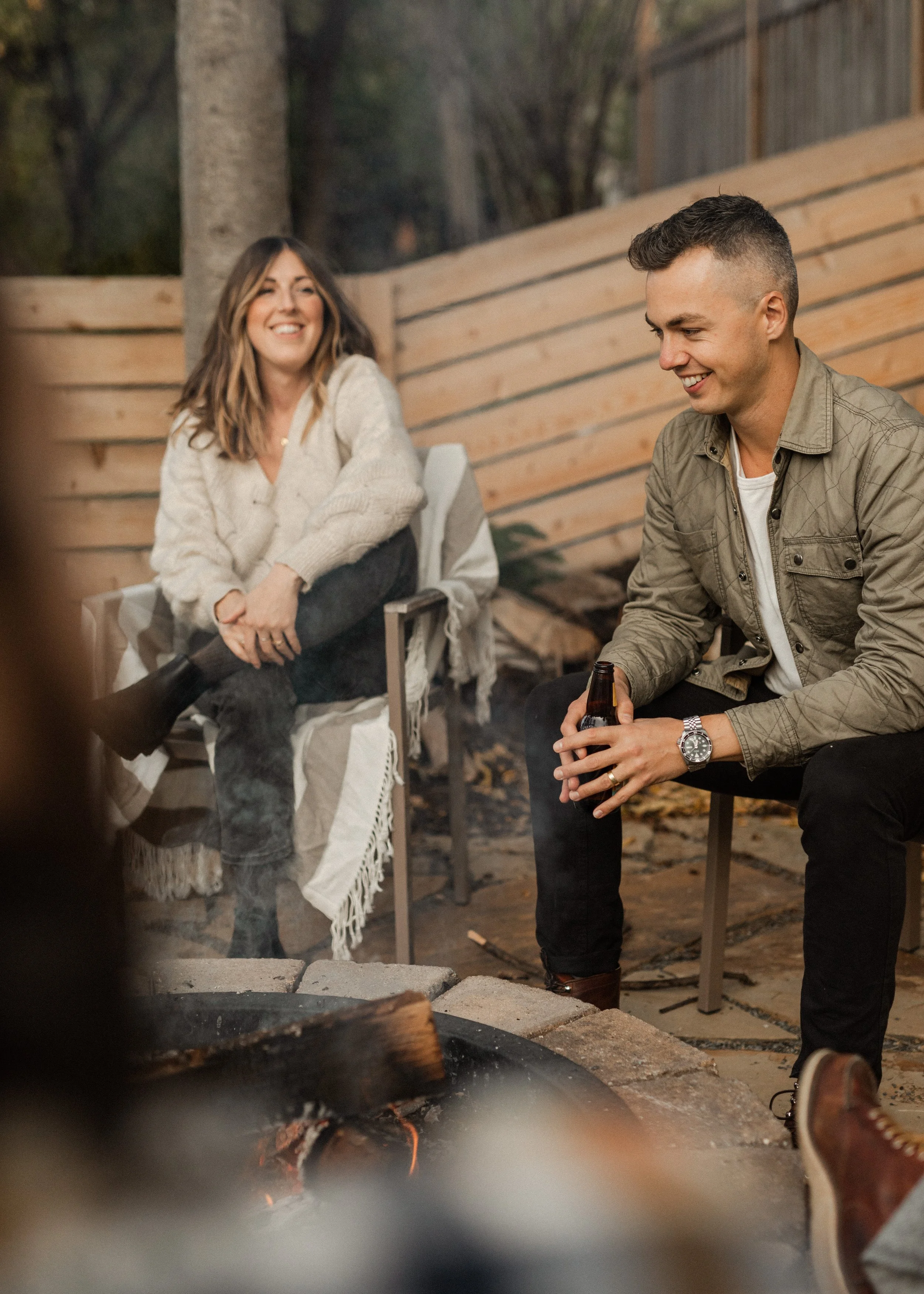 A man and a woman sit outdoors near a fire pit, smiling and chatting. The woman has long brown hair and wears a cream sweater, while the man has short hair and is dressed in a khaki jacket and dark pants, holding a beer bottle. They are sitting on ch