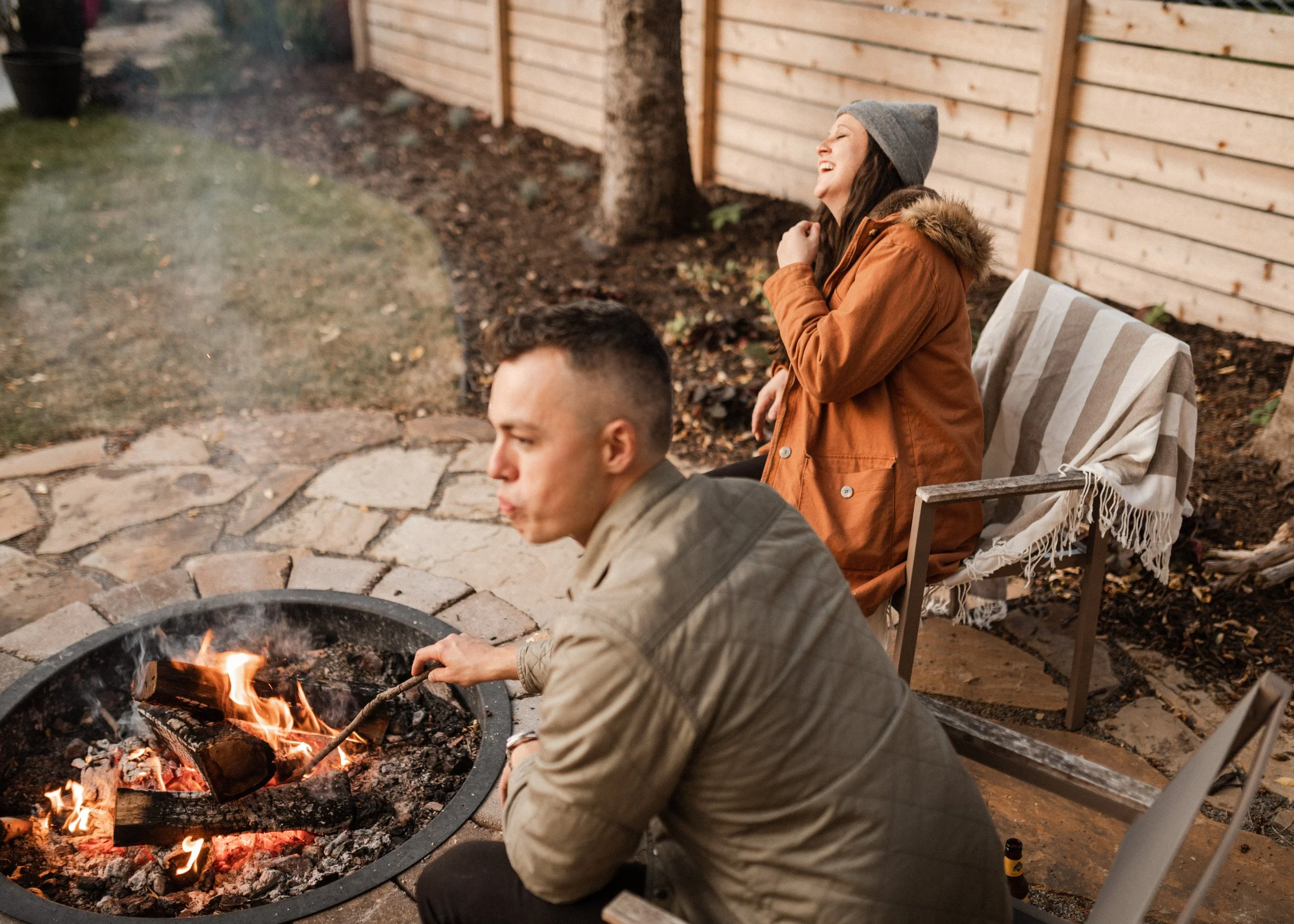 A woman sitting on a outdoor chair near a fire pit, laughing while wearing a gray beanie and an orange winter coat with a fur-lined hood. A man is in the foreground, tending the fire.
