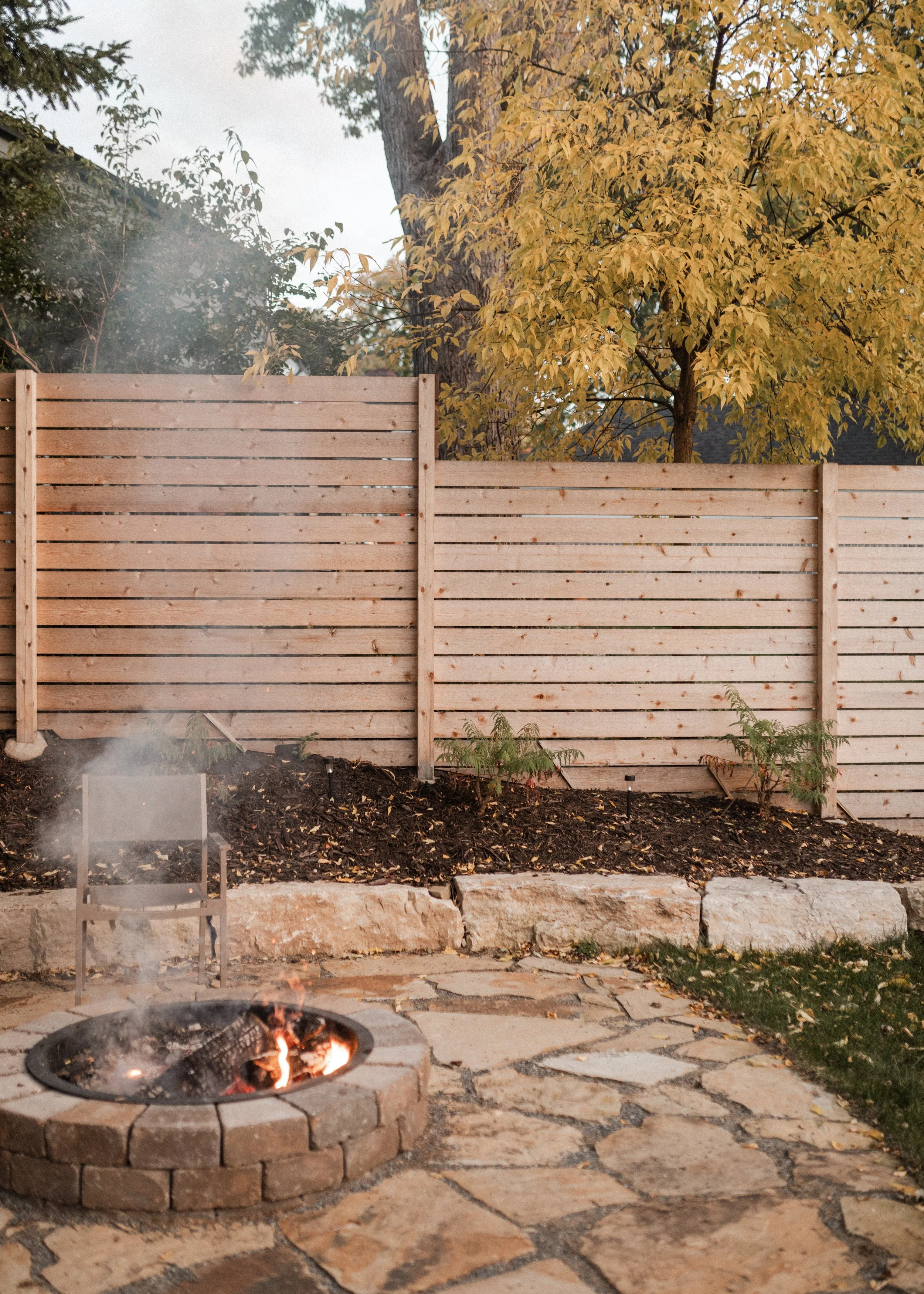 Backyard scene with a fire pit with a small fire, a metal chair nearby, a stone pathway, a wooden fence, a large tree with yellow autumn leaves, and some small plants and rocks on the ground.