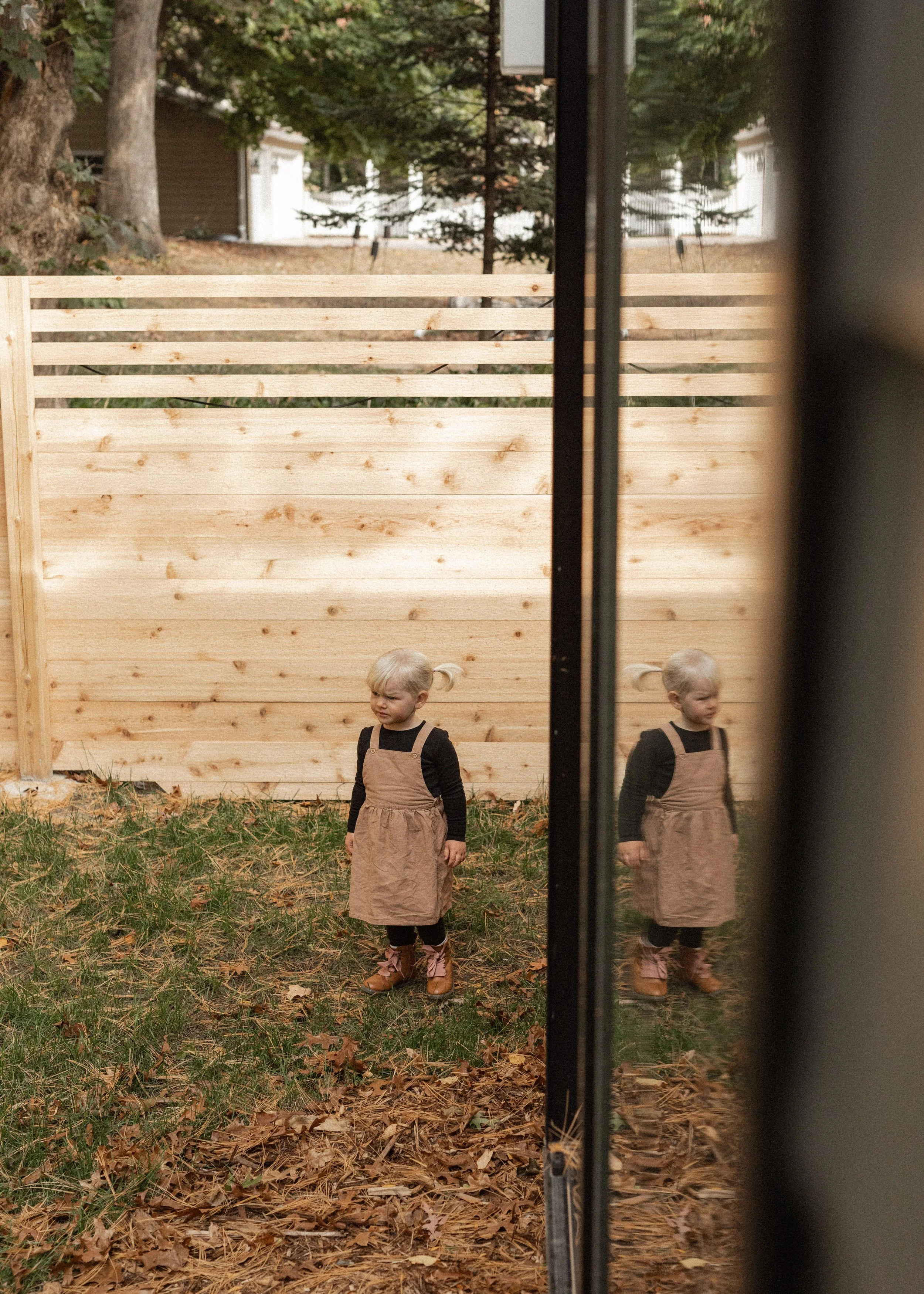 A young blonde girl with pigtails wearing a beige dress over a black long-sleeve shirt and brown boots, standing on a grassy area covered with fallen leaves in front of a wooden fence outdoors. Her reflection is seen in a glass door or window to the 