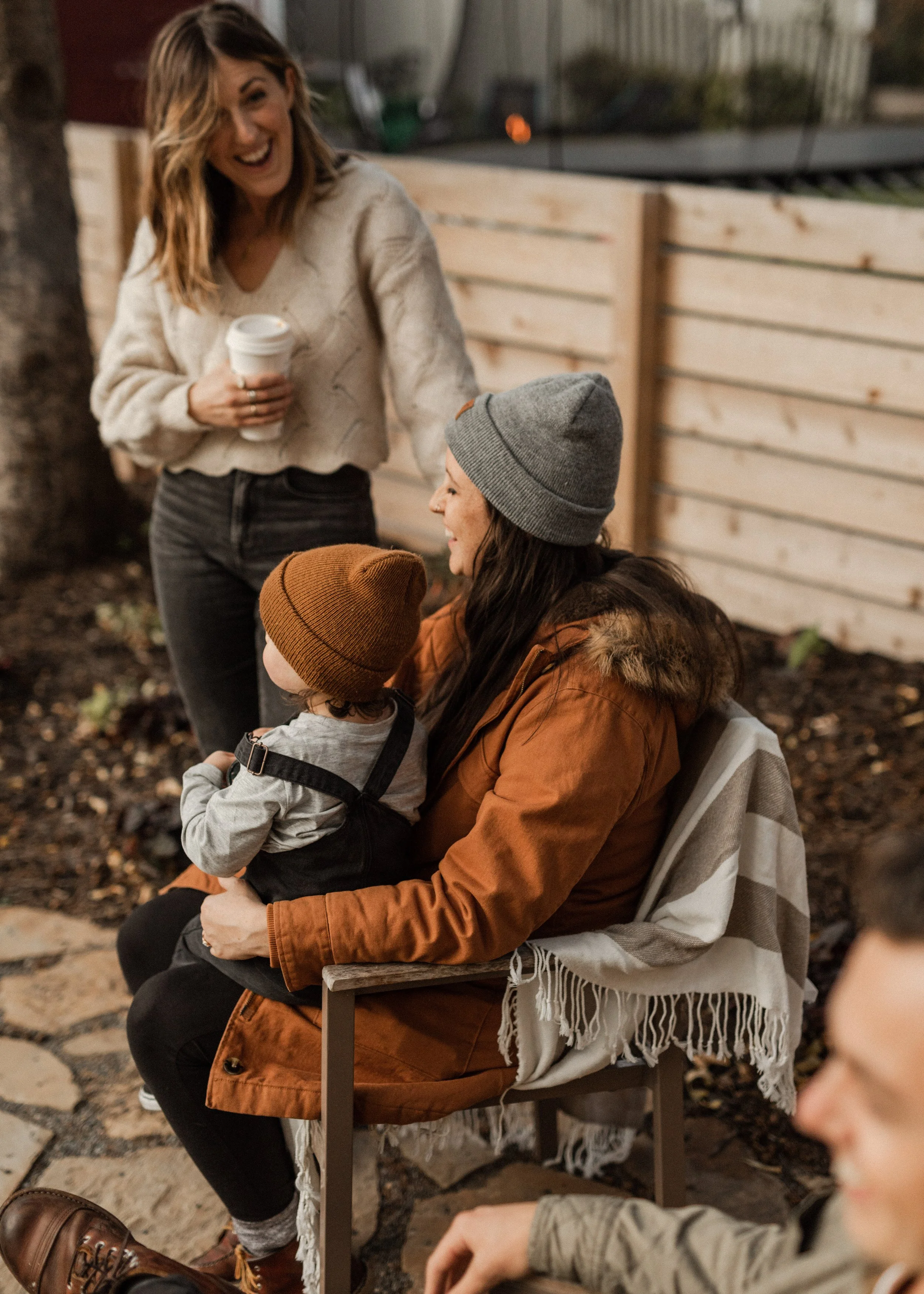 A woman standing outdoors, laughing and holding a coffee cup, engaging with others. Sitting on a bench nearby is a woman wearing a gray beanie and a woman in a brown jacket, with a small child in a tan beanie and gray sweater. The setting has a woode