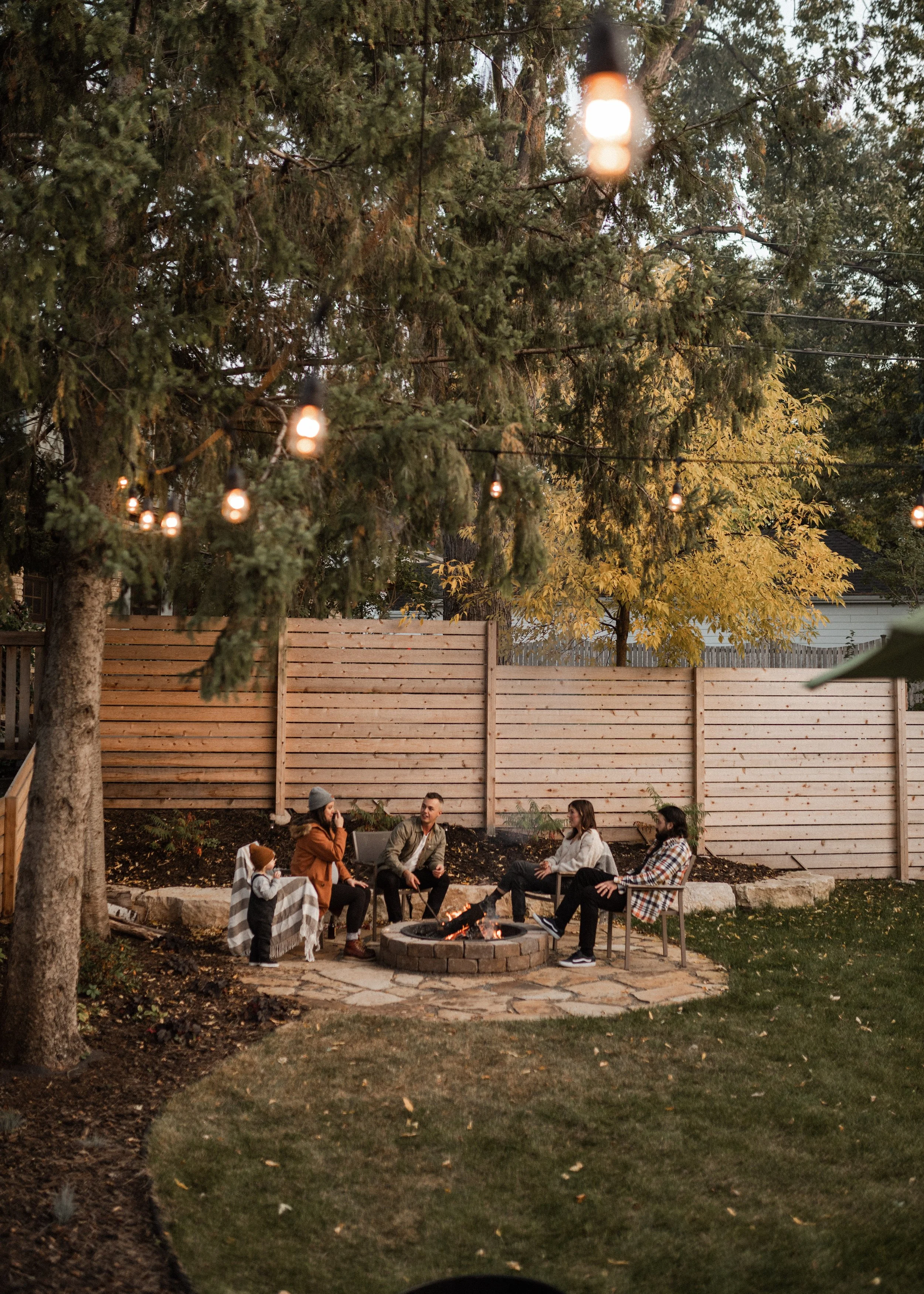 A group of seven people sitting around a fire pit in a backyard, with a string of outdoor lights overhead and a wooden fence in the background, during the evening.