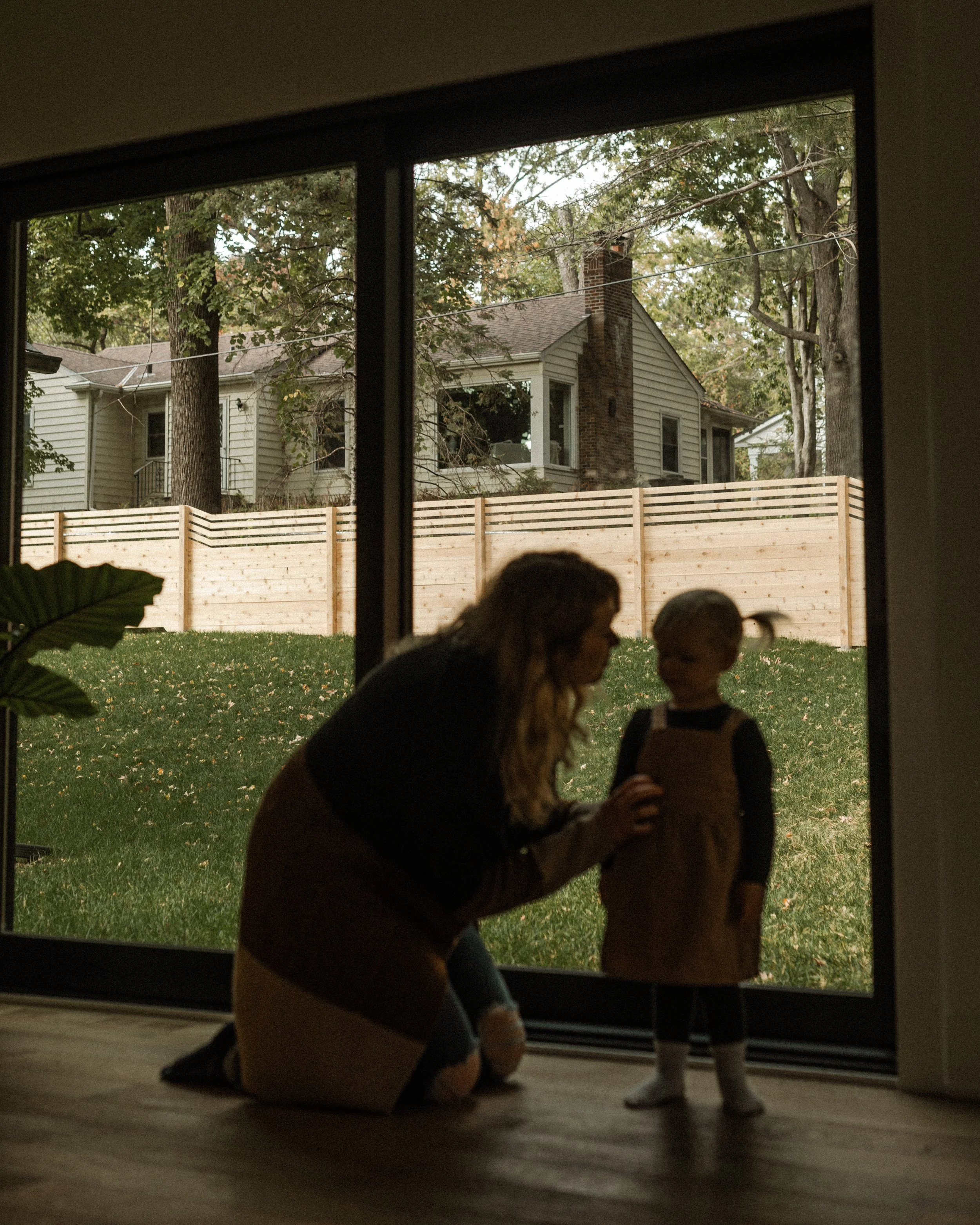 A woman kneeling and a girl talking through a window, with an outdoor yard and trees visible in the background.