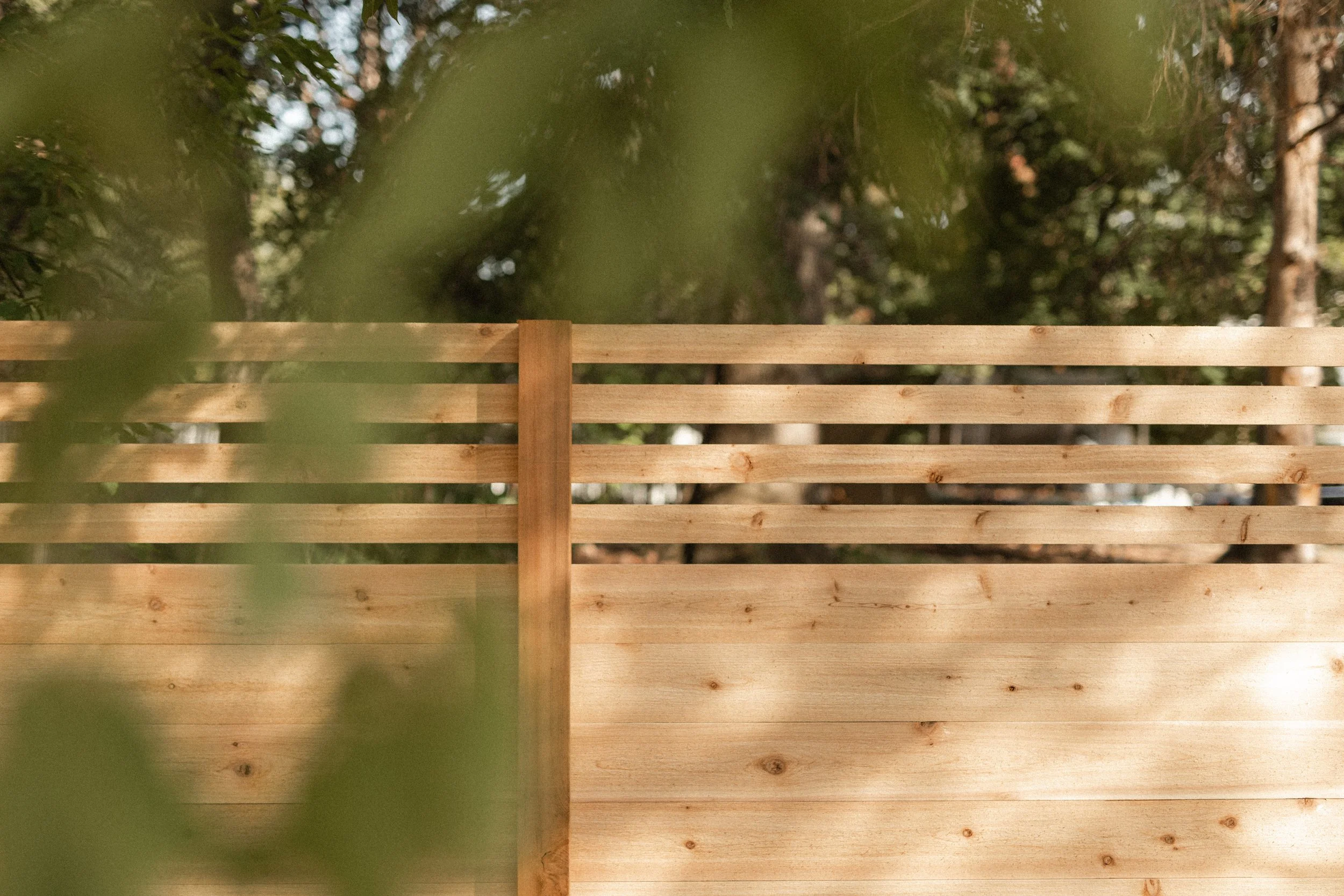 A wooden fence with horizontal slats in a natural wood finish, set against a background of blurred trees and foliage in a forest setting.
