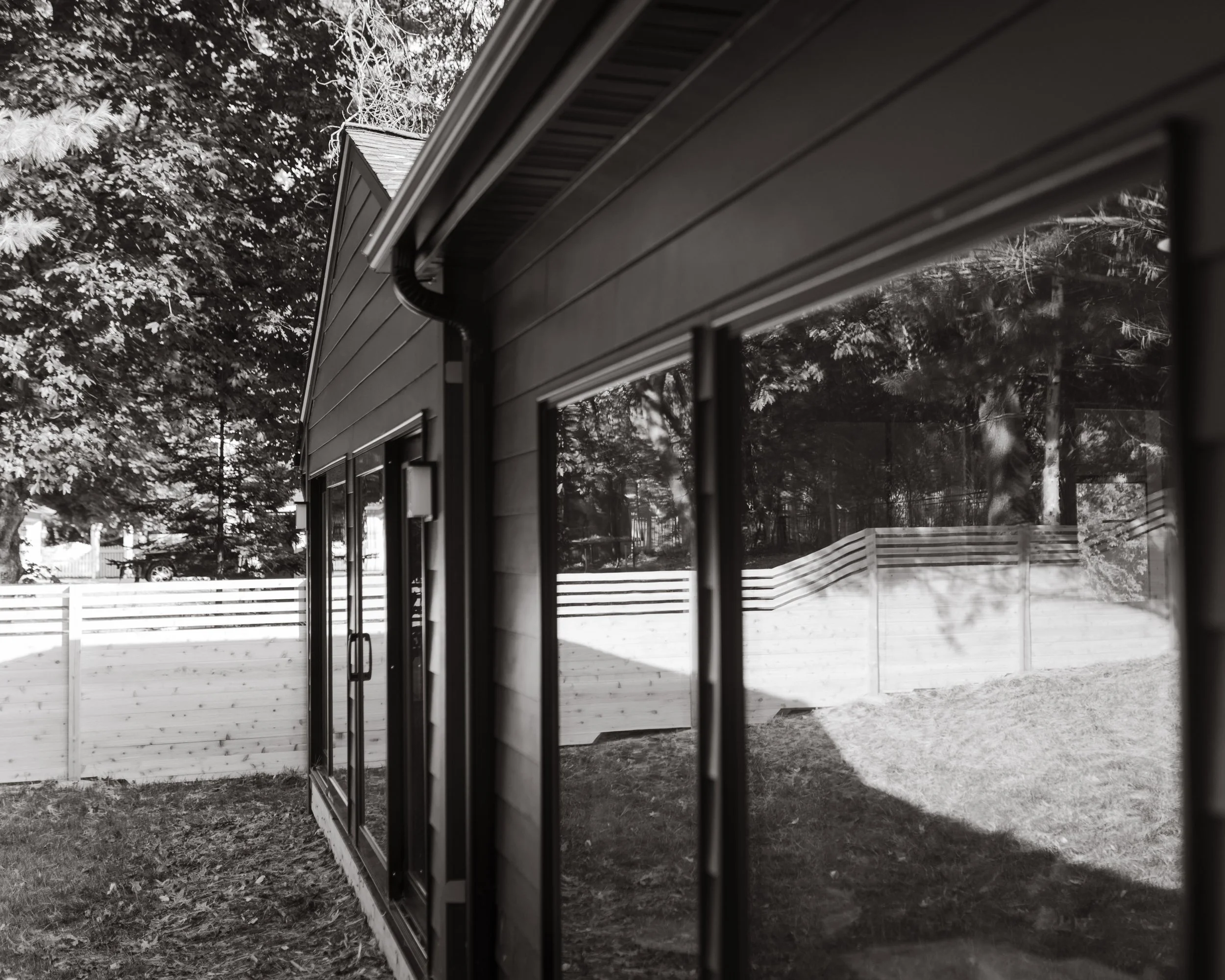 Black and white photo of the exterior of a house with large glass windows, a gutter, and a wooden fence in the yard with trees in the background.