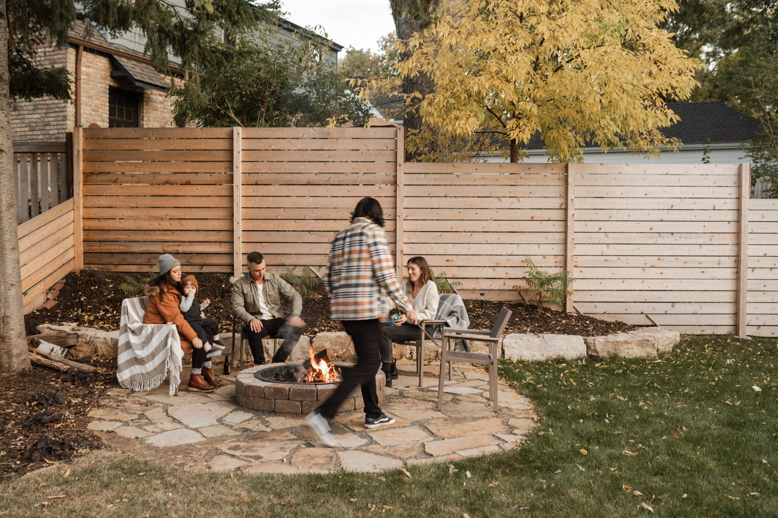 A group of people sitting around a fire pit in a backyard, with some smoking and talking, while a woman in a plaid shirt stands nearby. There is a wooden fence, trees with yellow leaves, and a house in the background.