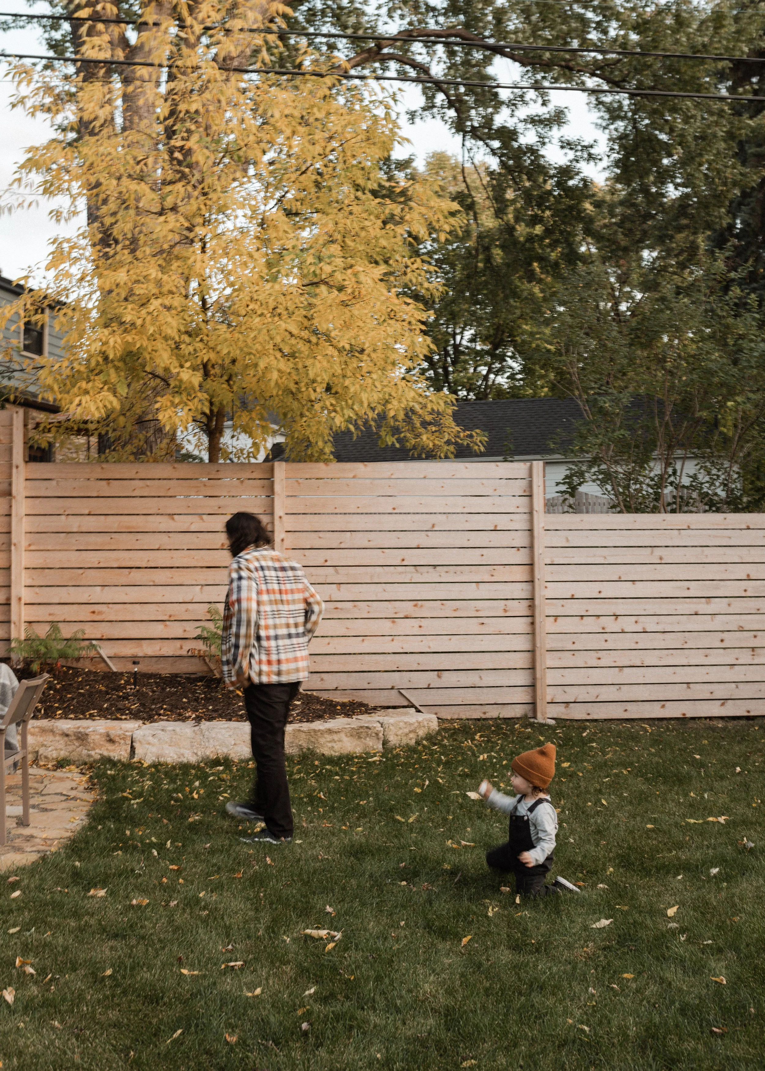 A backyard scene with a wooden fence, trees with yellow and green leaves, a child on their knees in the grass wearing a brown hat, gray and black clothing, and a water bottle in hand, and an adult walking past them.