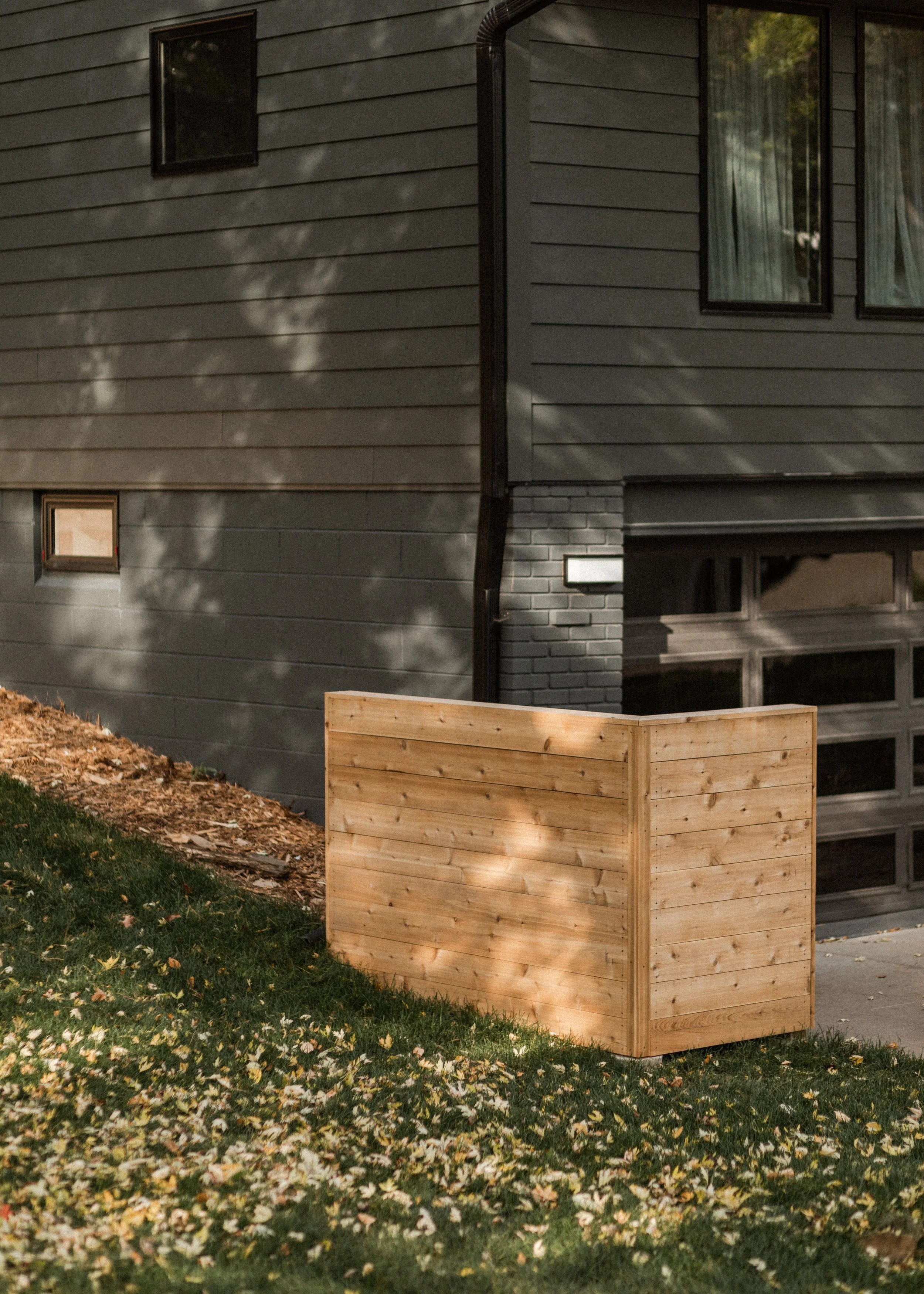 A house with gray horizontal siding and black-framed windows. A small window is near the ground, and larger windows are higher up. A black downspout runs along the corner of the house. A wooden planter box is in the foreground on a grassy yard with f