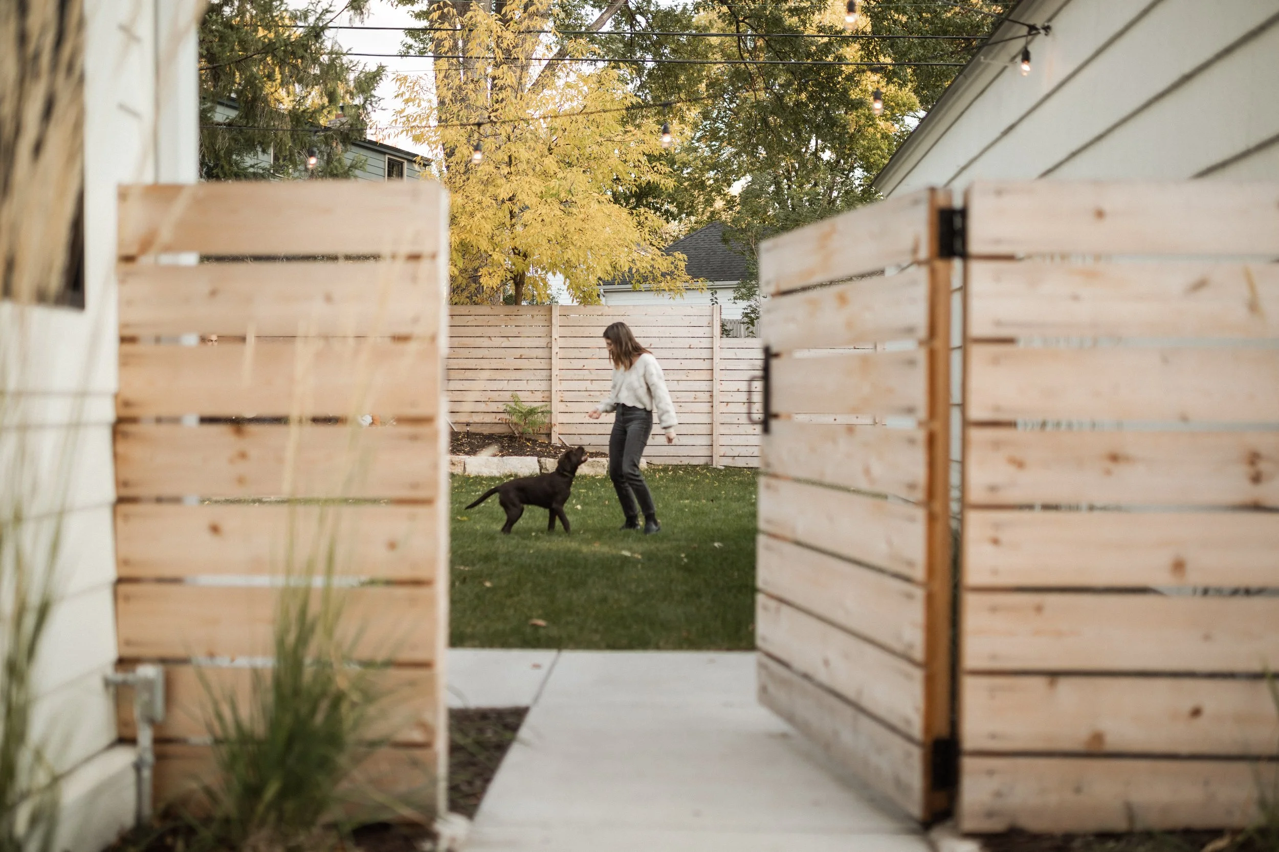 A woman playing with a black dog in a backyard, framed by wooden fences and lit string lights, with trees and a house in the background.