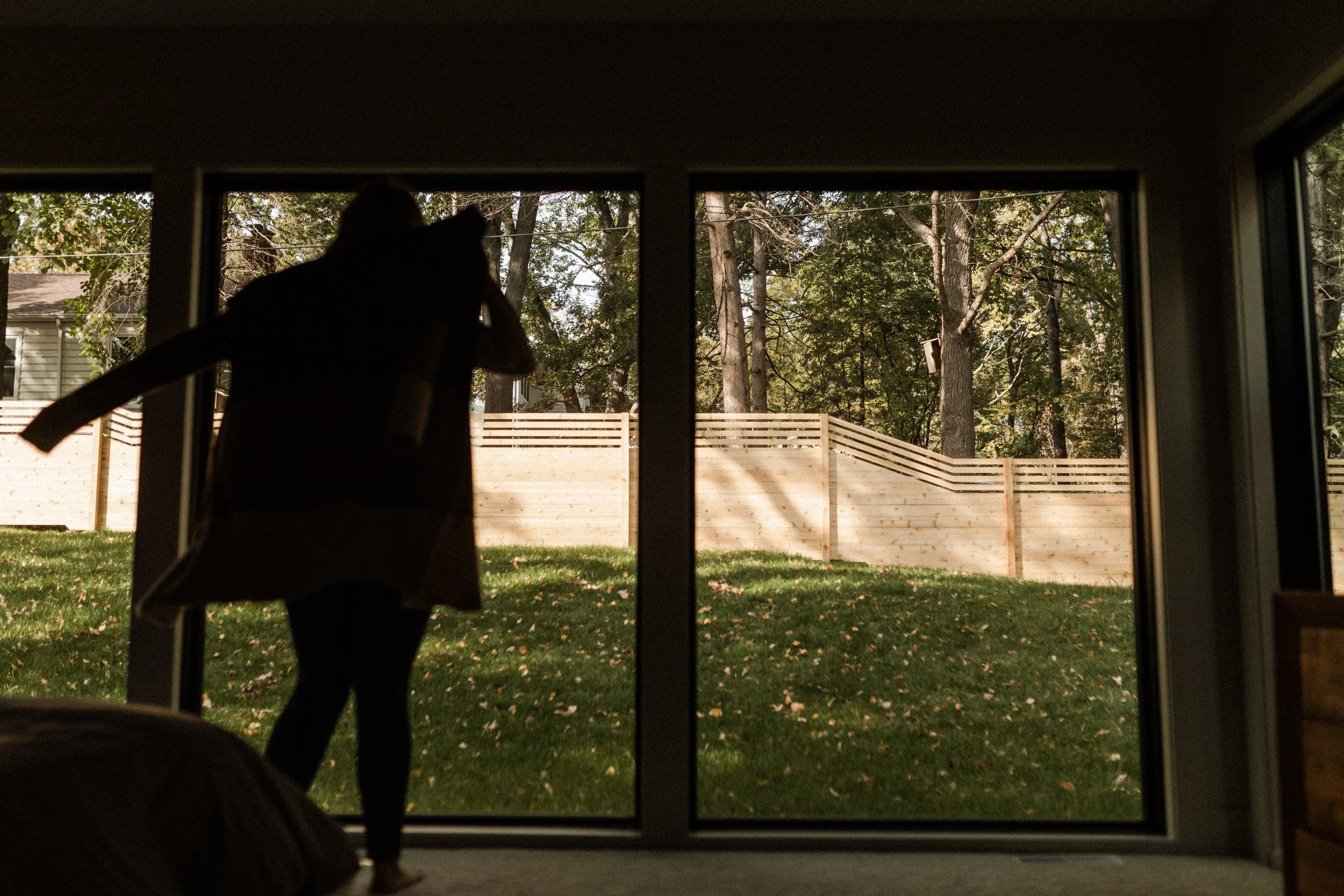 A person standing indoors near a large window, looking outside at a backyard with trees, a wooden fence, and grass.