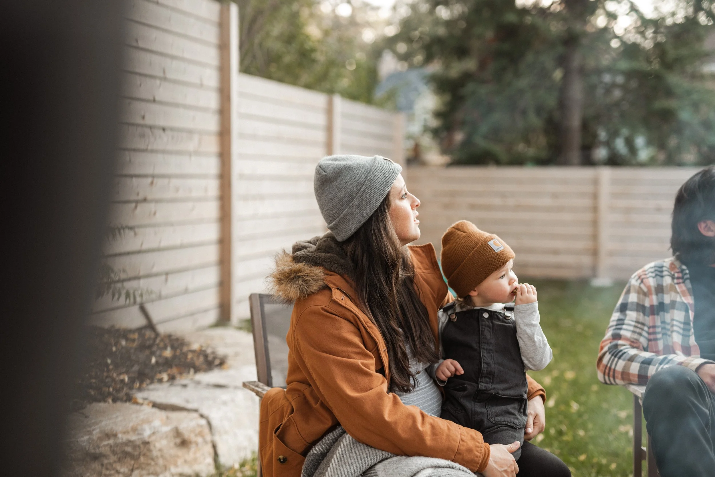A woman with long brown hair wearing a gray beanie and an orange jacket sitting outdoors with two young children, a girl and a boy, during fall. The girl is wearing a mustard-colored beanie and a black jacket, and the boy is in a plaid shirt. They ar