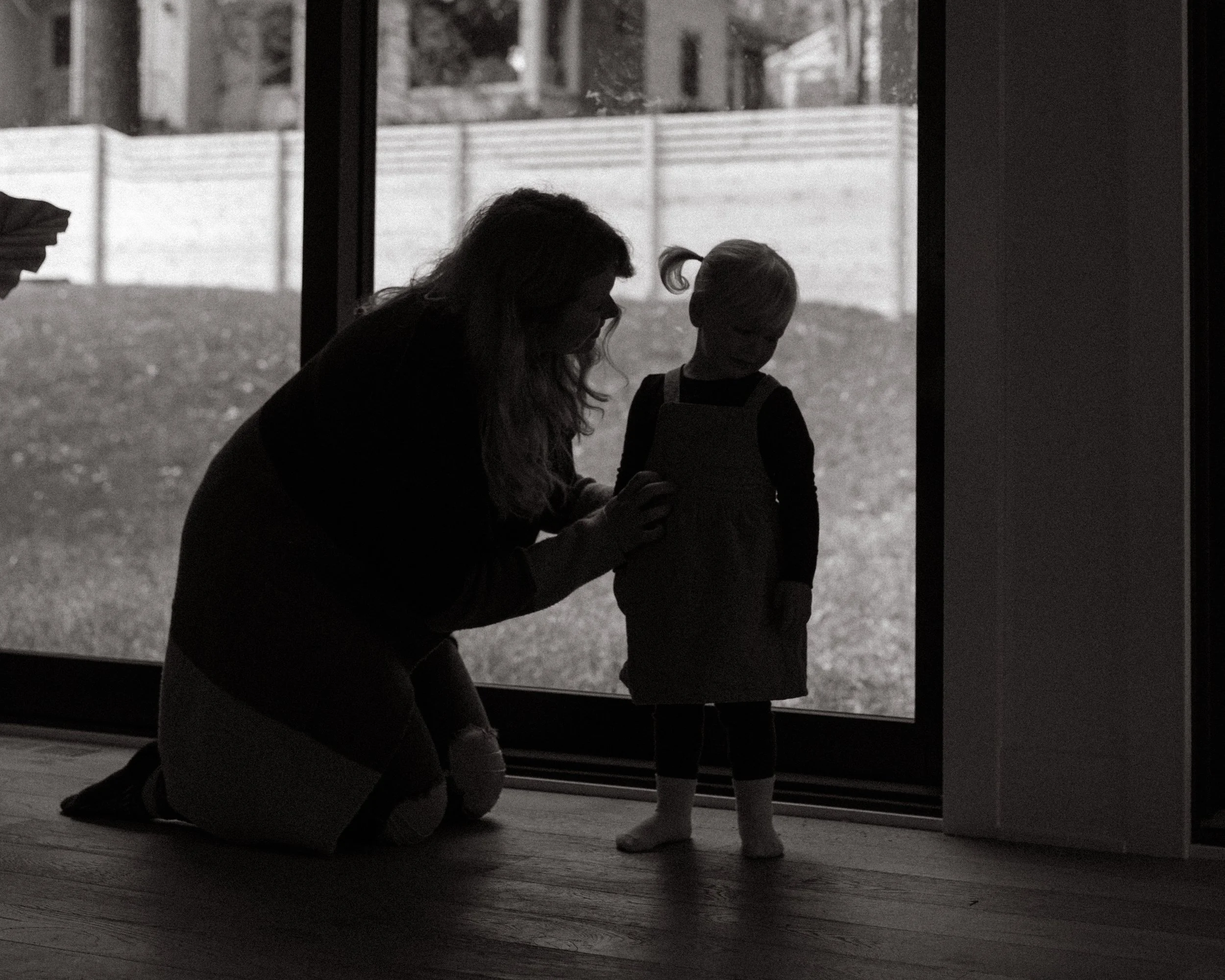 A black and white photo of a woman kneeling on the floor, reaching out to a young child, who is standing and facing her. They are indoors next to large windows with an outdoor yard and fencing visible in the background.