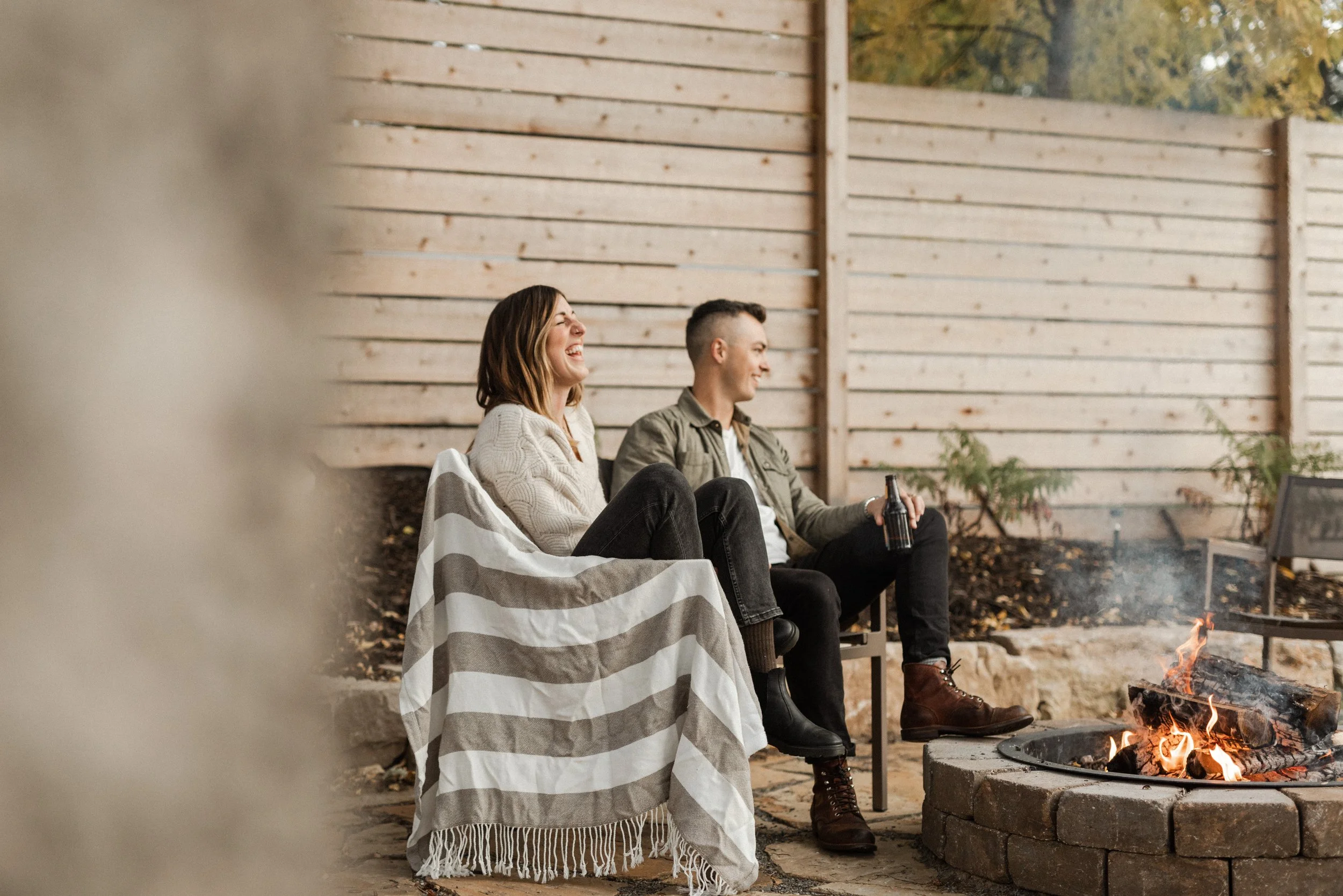 Two people sitting by a fire pit outdoors during fall, one person laughing and the other smiling. They are sitting on chairs, with a blanket draped over the person's lap, and the ground is covered with fallen leaves. A wooden fence is in the backgrou