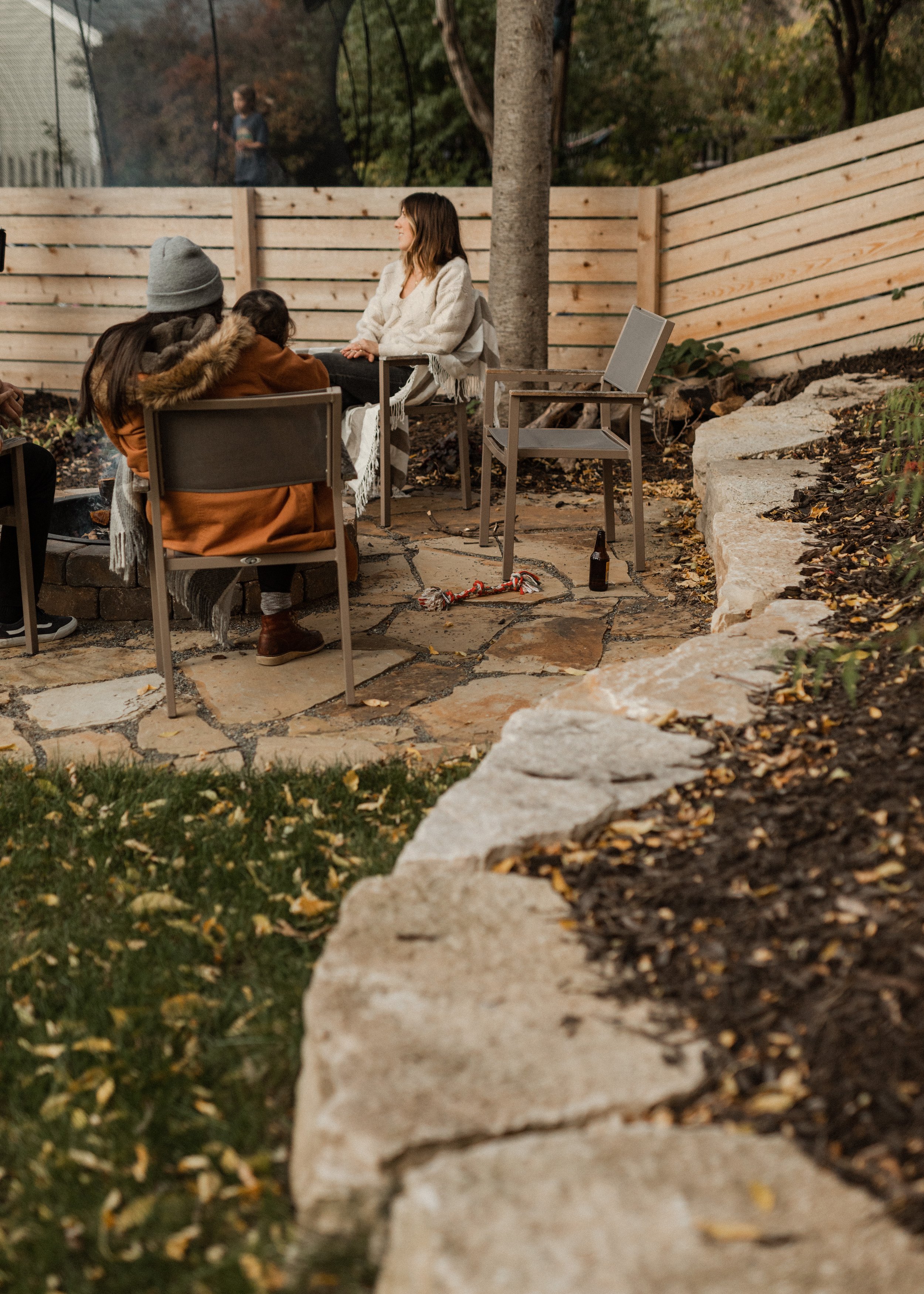 Three people sitting around a fire pit in a backyard, engaging in conversation, with a wooden fence and a tree in the background.