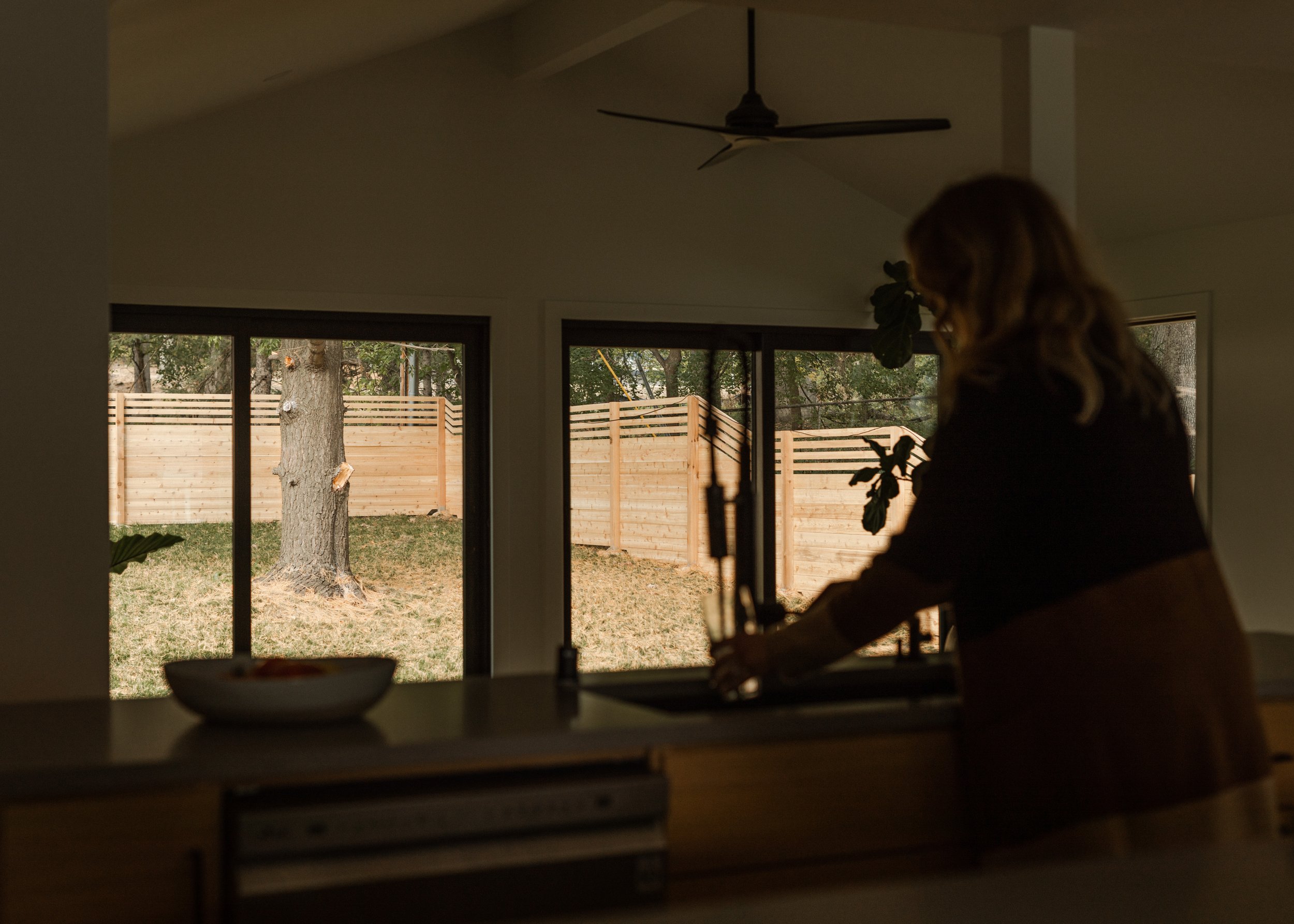 A person with long hair in a dark top is inside and appears to be working on something, with a view of a backyard through large glass sliding doors. The backyard has a tree, a wooden fence, and some outdoor construction or gardening equipment. The ph