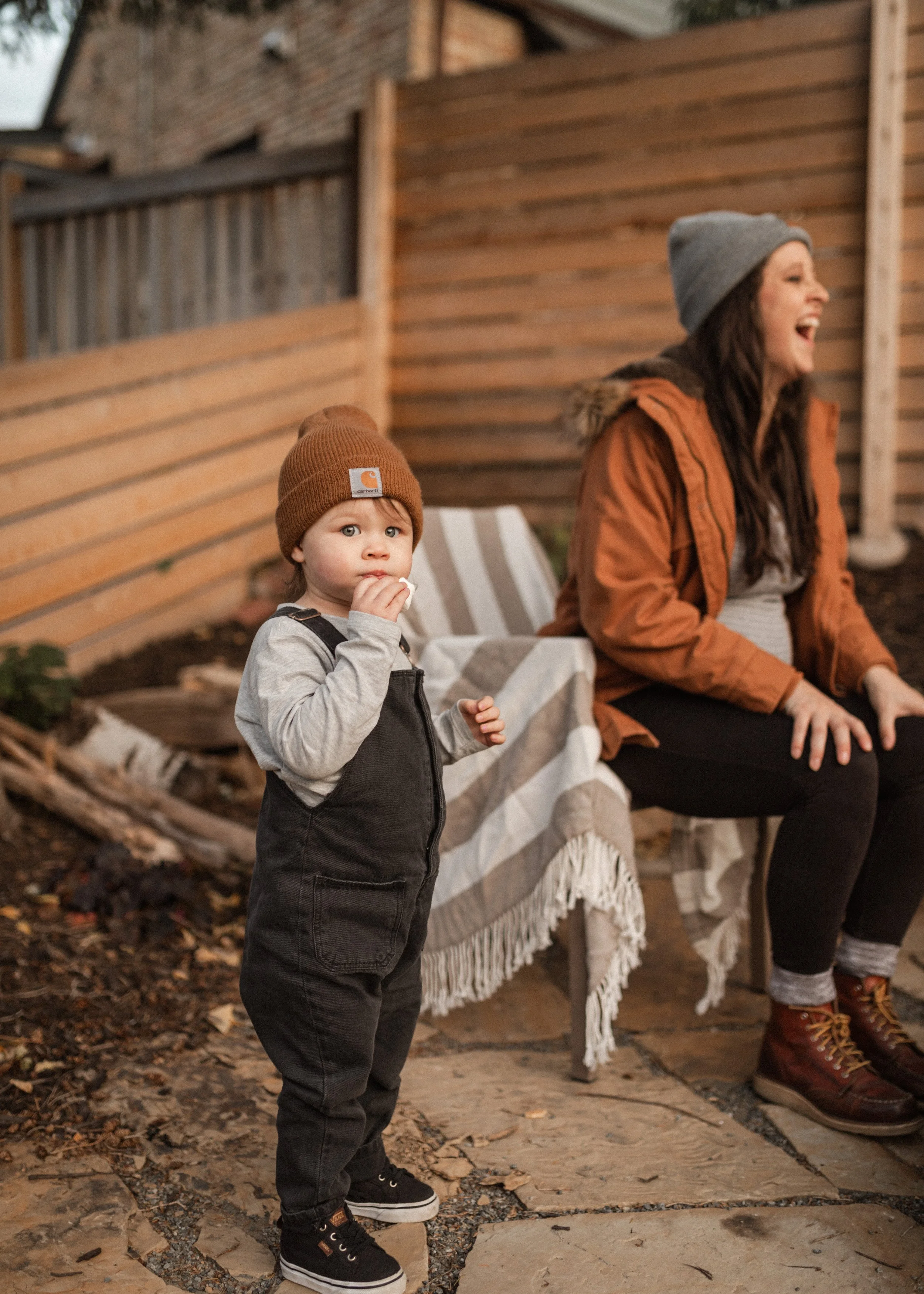 A young boy in a brown beanie, grey shirt, and black overalls stands outdoors on a stone and leaf-covered ground, looking at the camera and eating a snack, while a woman with long dark hair, wearing a grey beanie, orange jacket, and black pants, laug