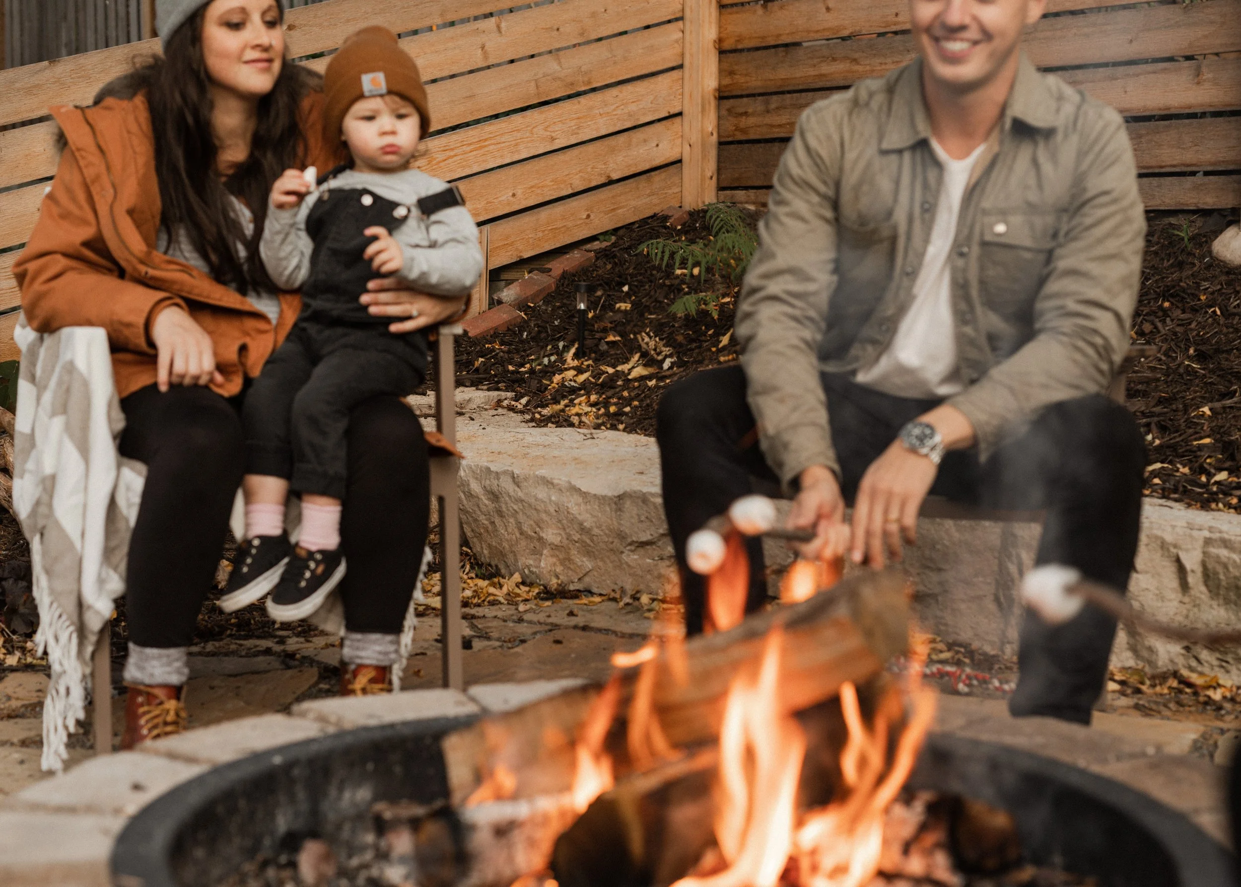 A family sitting around a firepit in a backyard, enjoying a night outdoors. The woman holds a young child on her lap, and a man is sitting nearby. The setting is enclosed by a wooden fence, with mulch and small plants around the fire.