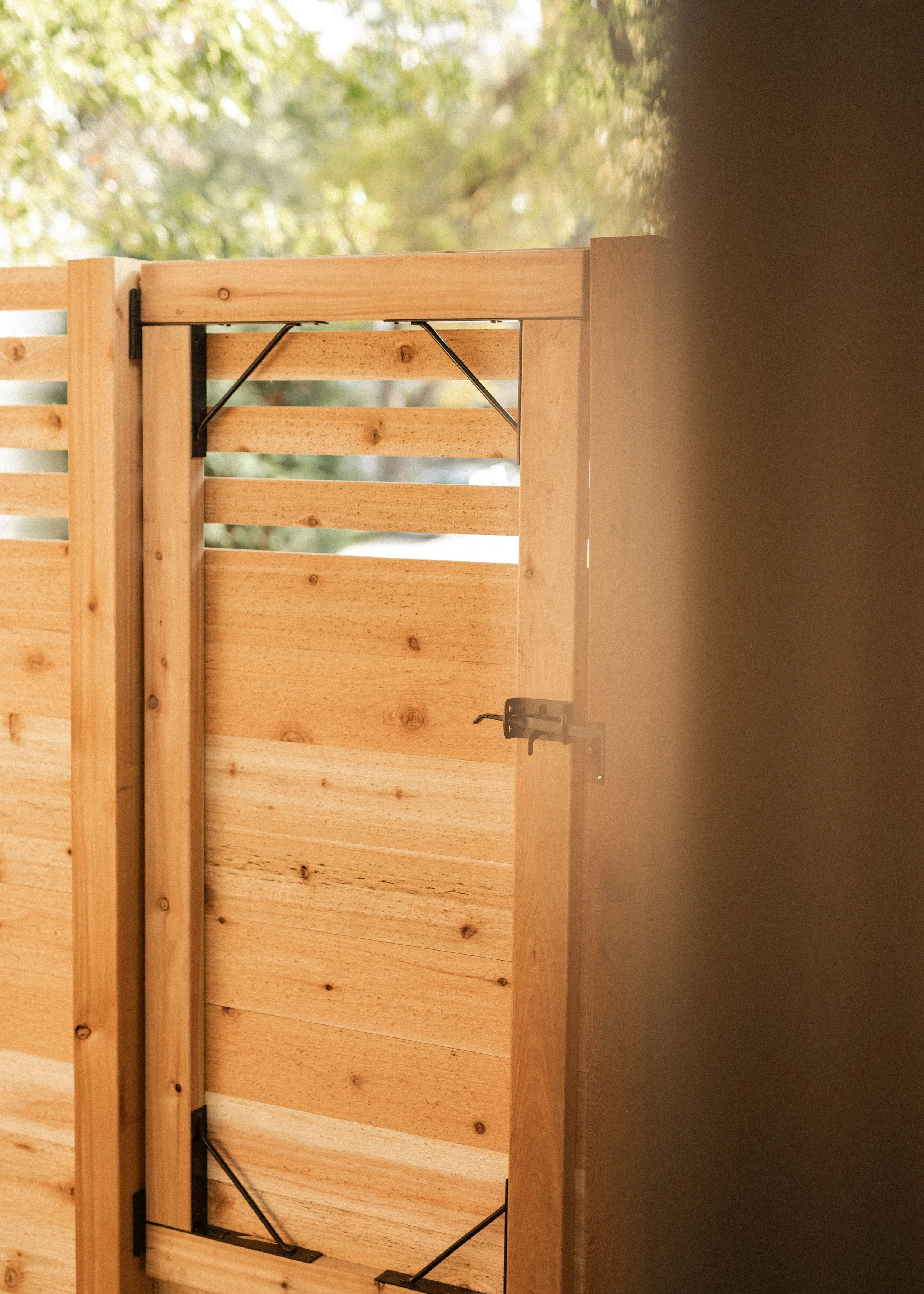 A wooden slatted gate with black hinges and diagonal support bars, set against a background of blurred greenery.