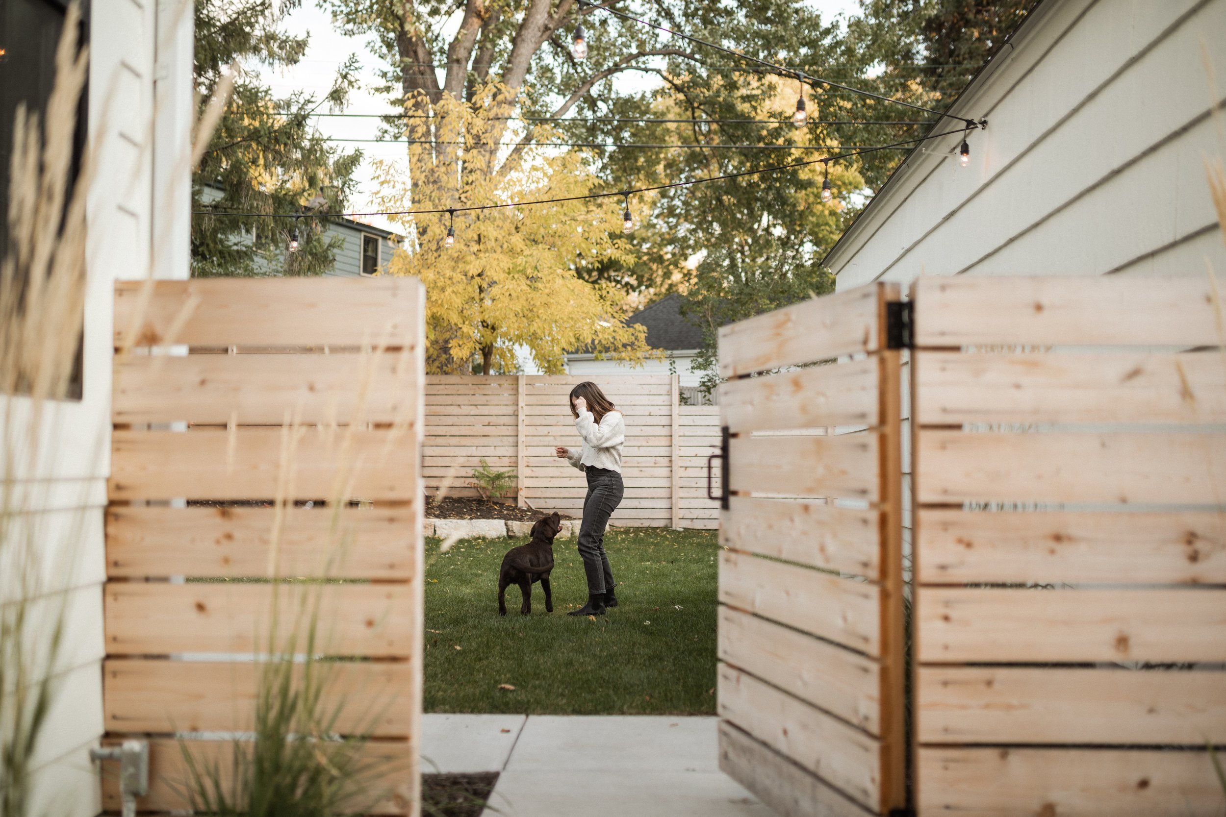 A woman standing outside in a backyard with a black dog, surrounded by a wooden fence, trees with yellow and green leaves, and string lights hanging overhead.