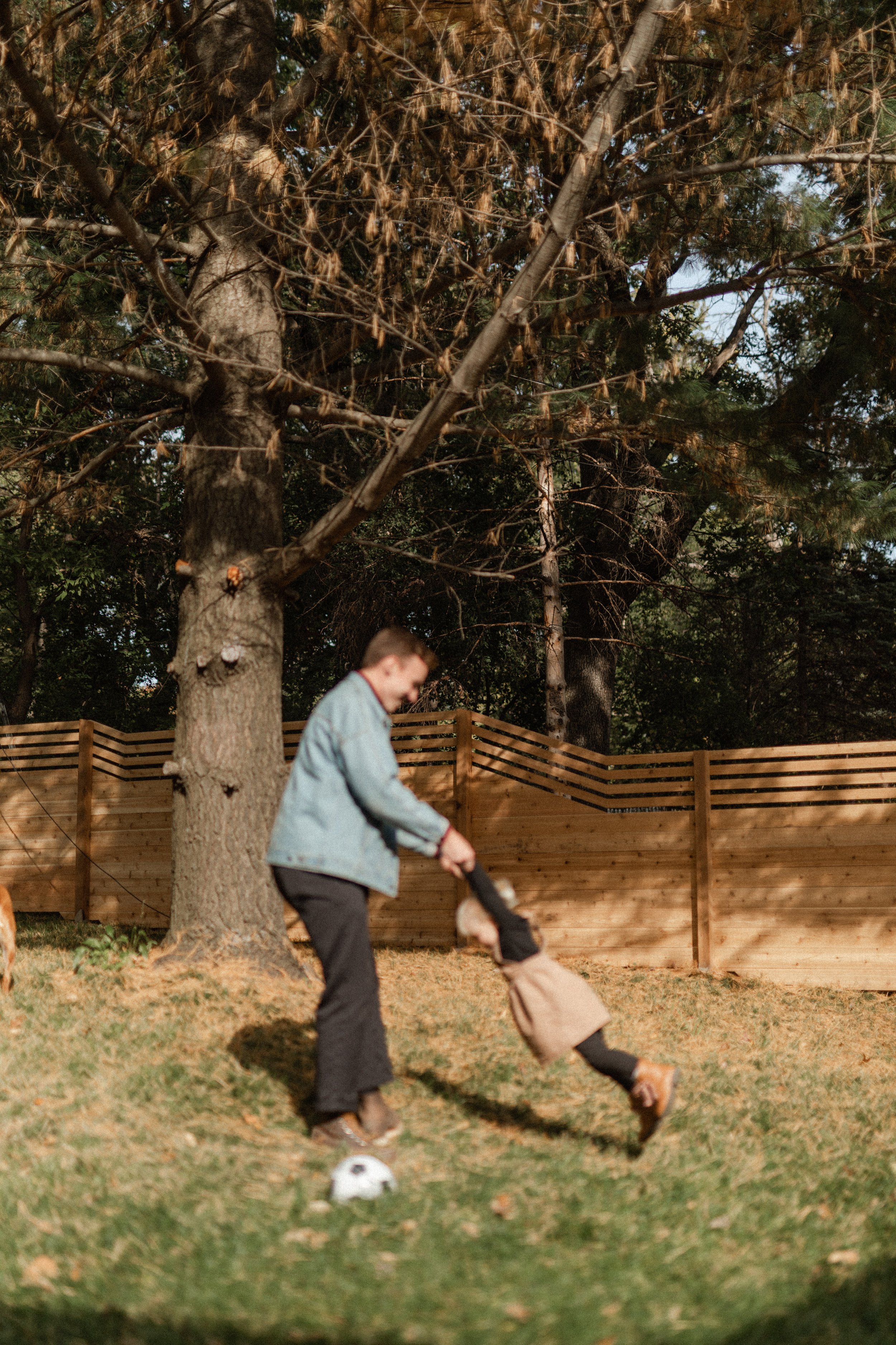 A person wearing a light blue jacket and black pants is playing soccer in a backyard. They are kicking a white soccer ball. The yard has a grassy area, a wooden fence, and trees with some brown leaves.