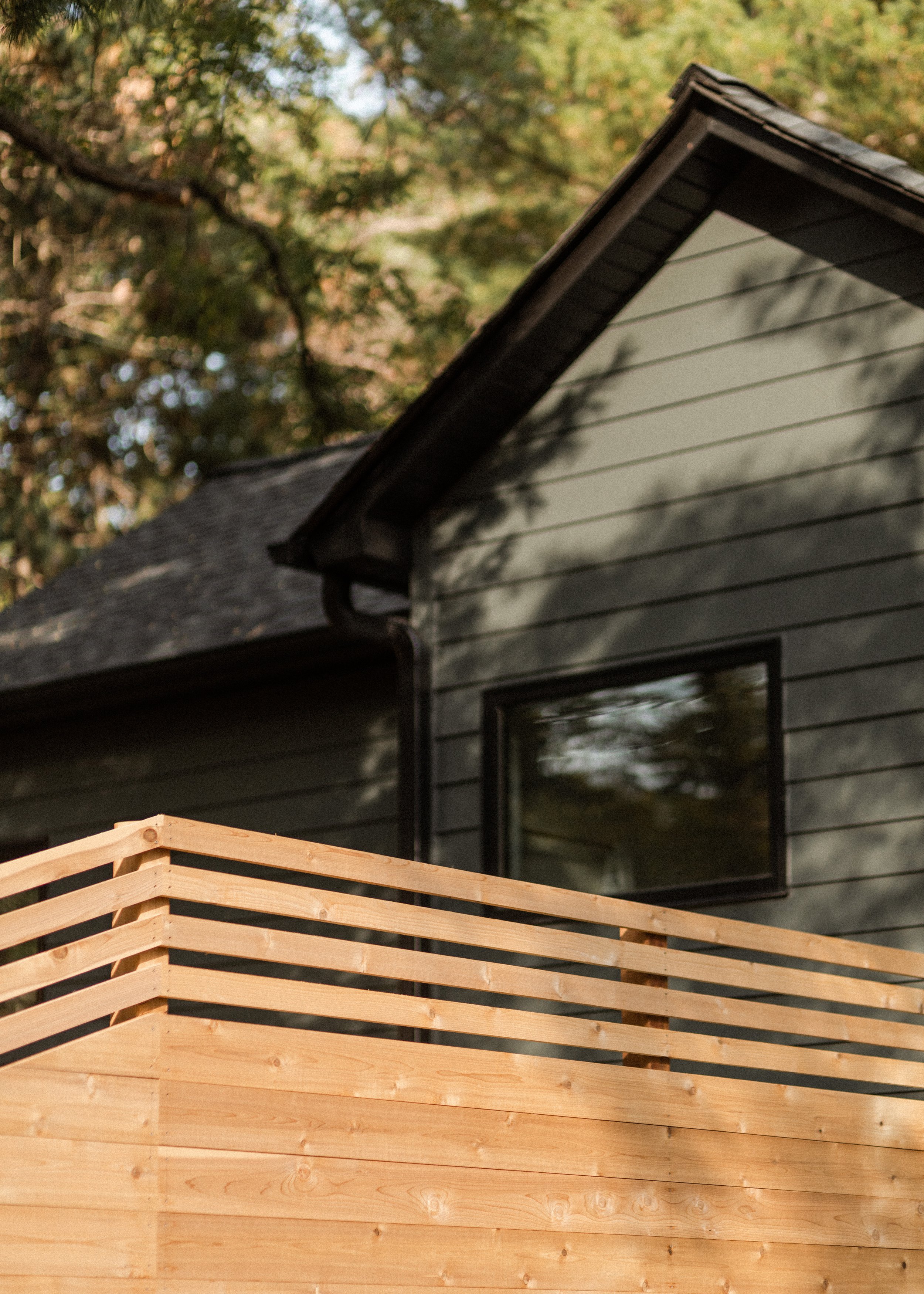 A house with gray wooden siding and black window frames, surrounded by trees, with a newly constructed light wood fence in the foreground.