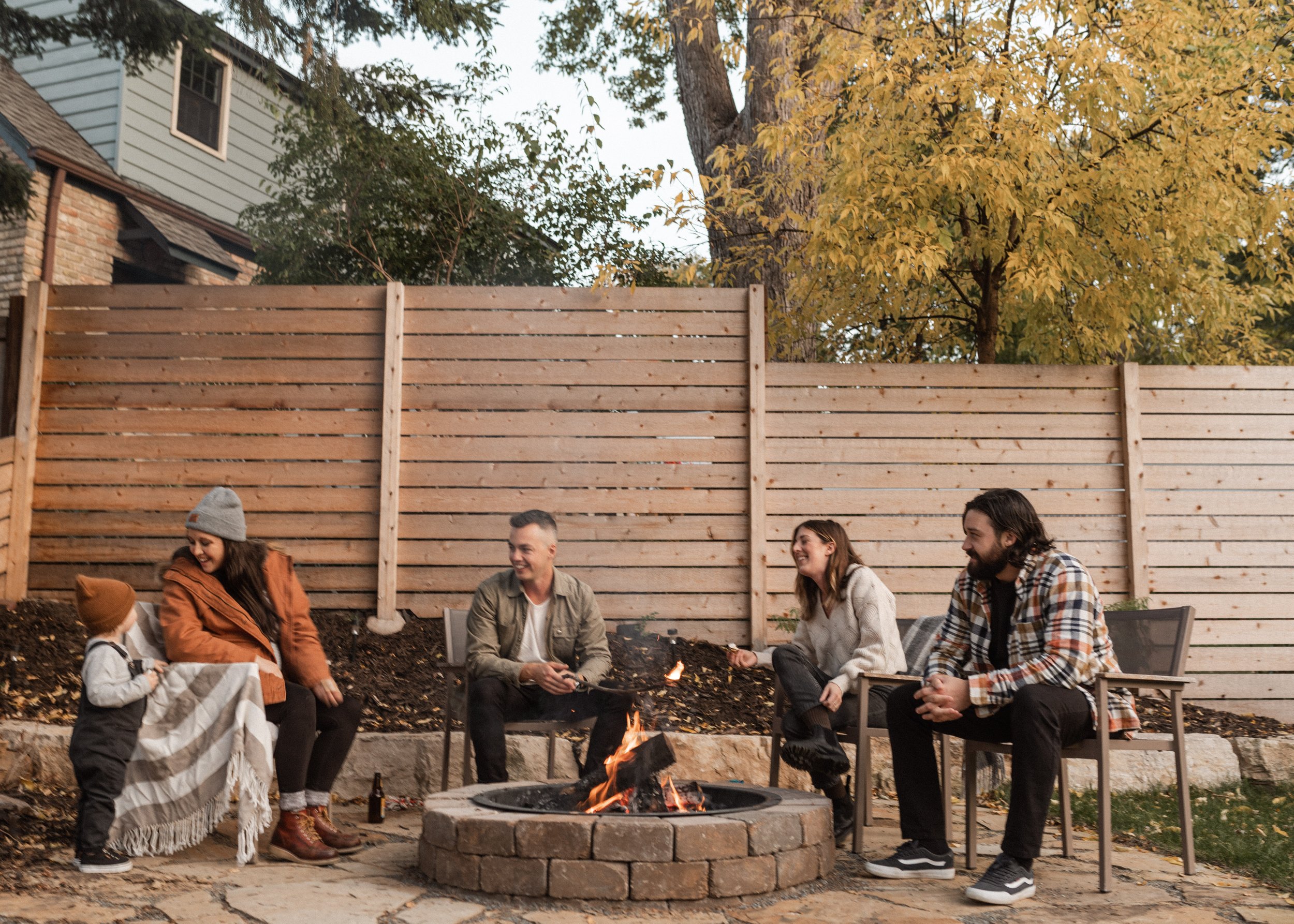 People sitting around a fire pit in a backyard, engaged in conversation and smiling, with a wooden fence, autumn trees, and a house in the background.