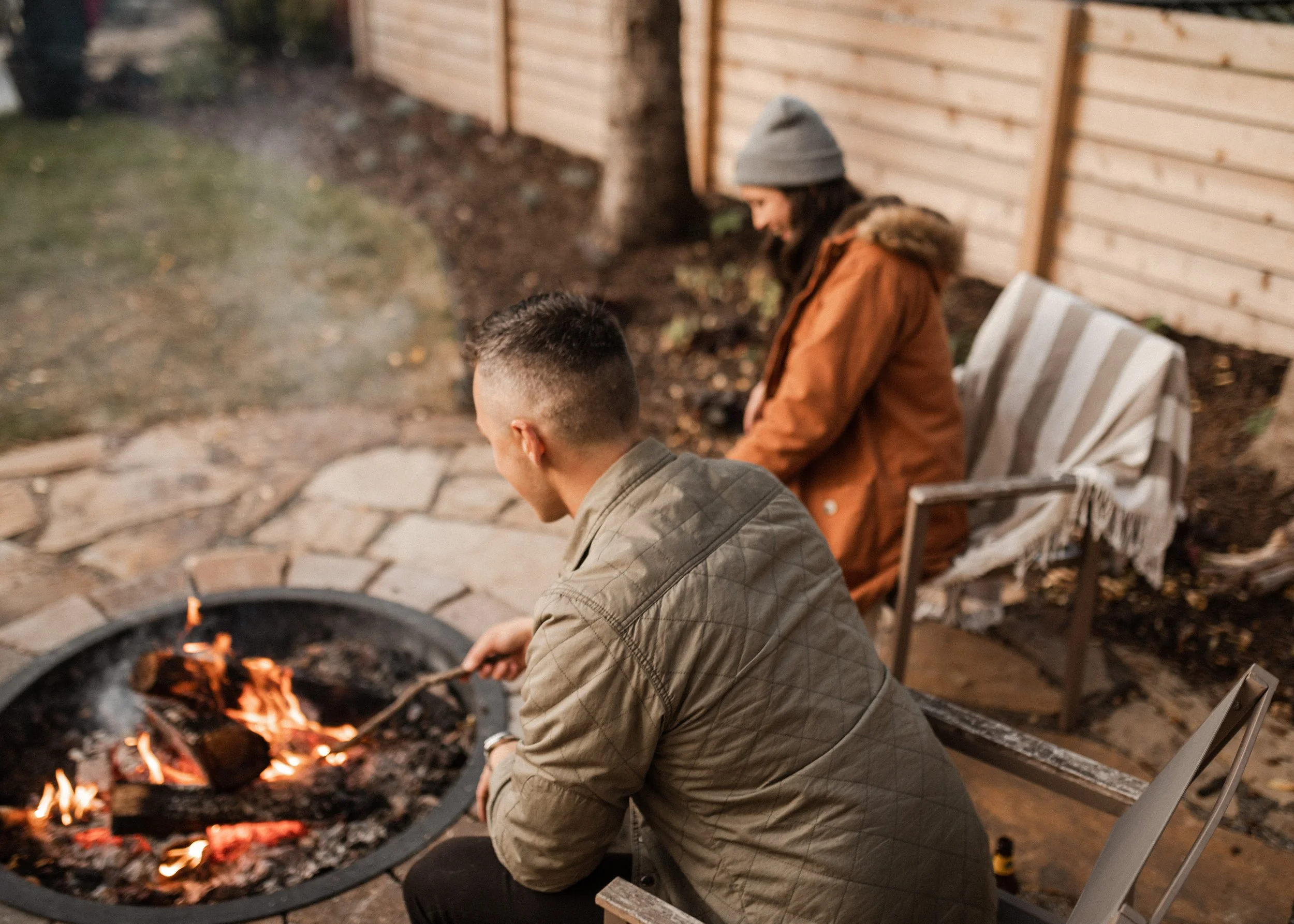 A person with short hair and a man with long hair wearing a gray beanie and brown jacket sitting by a fire pit outside next to a white bench and wooden fence.