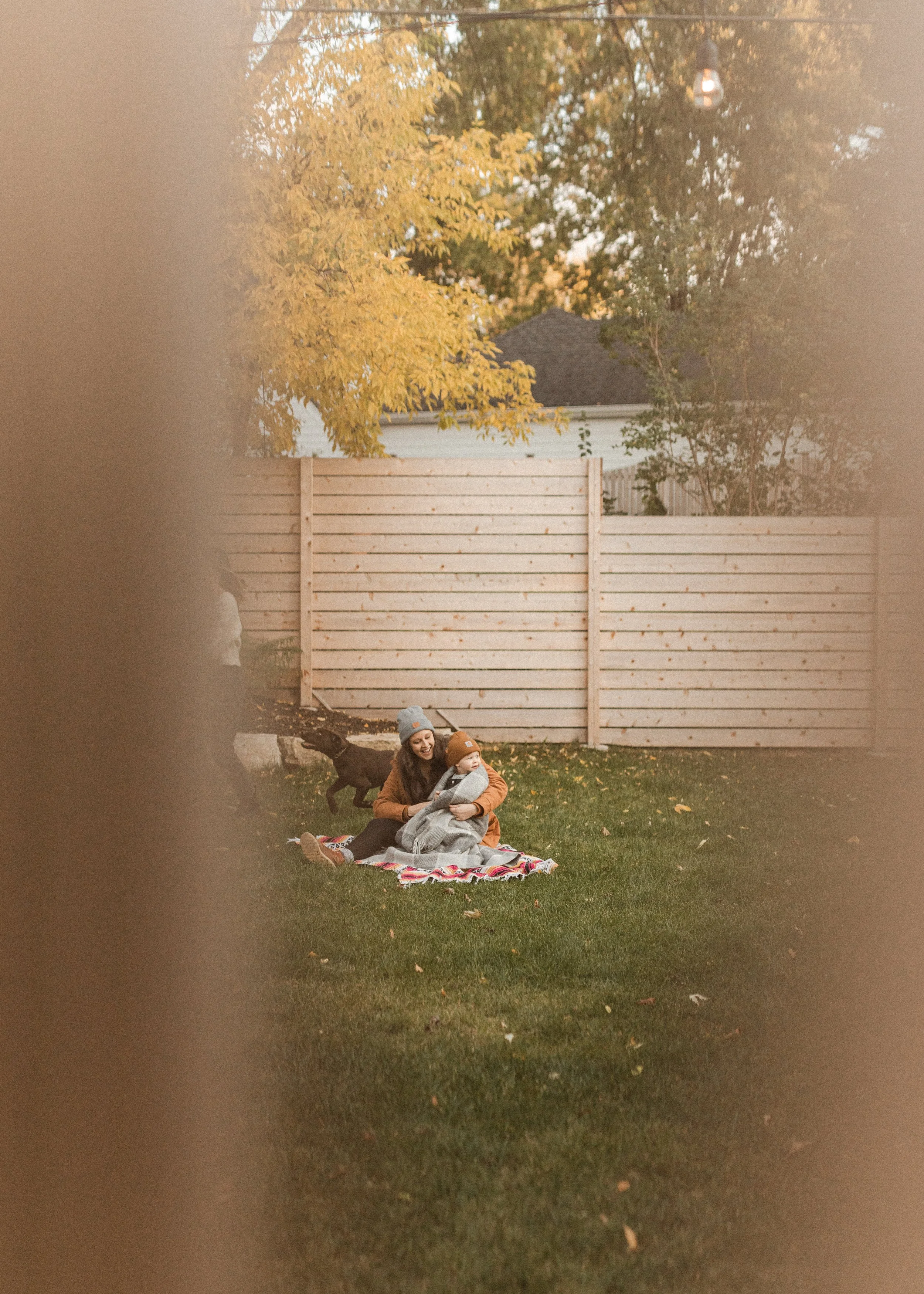 A woman and a young child sitting on a blanket on grass in a backyard, surrounded by trees with autumn foliage, with a wooden fence and a house in the background. The woman is wearing a gray beanie and a brown jacket, while the child wears an orange 