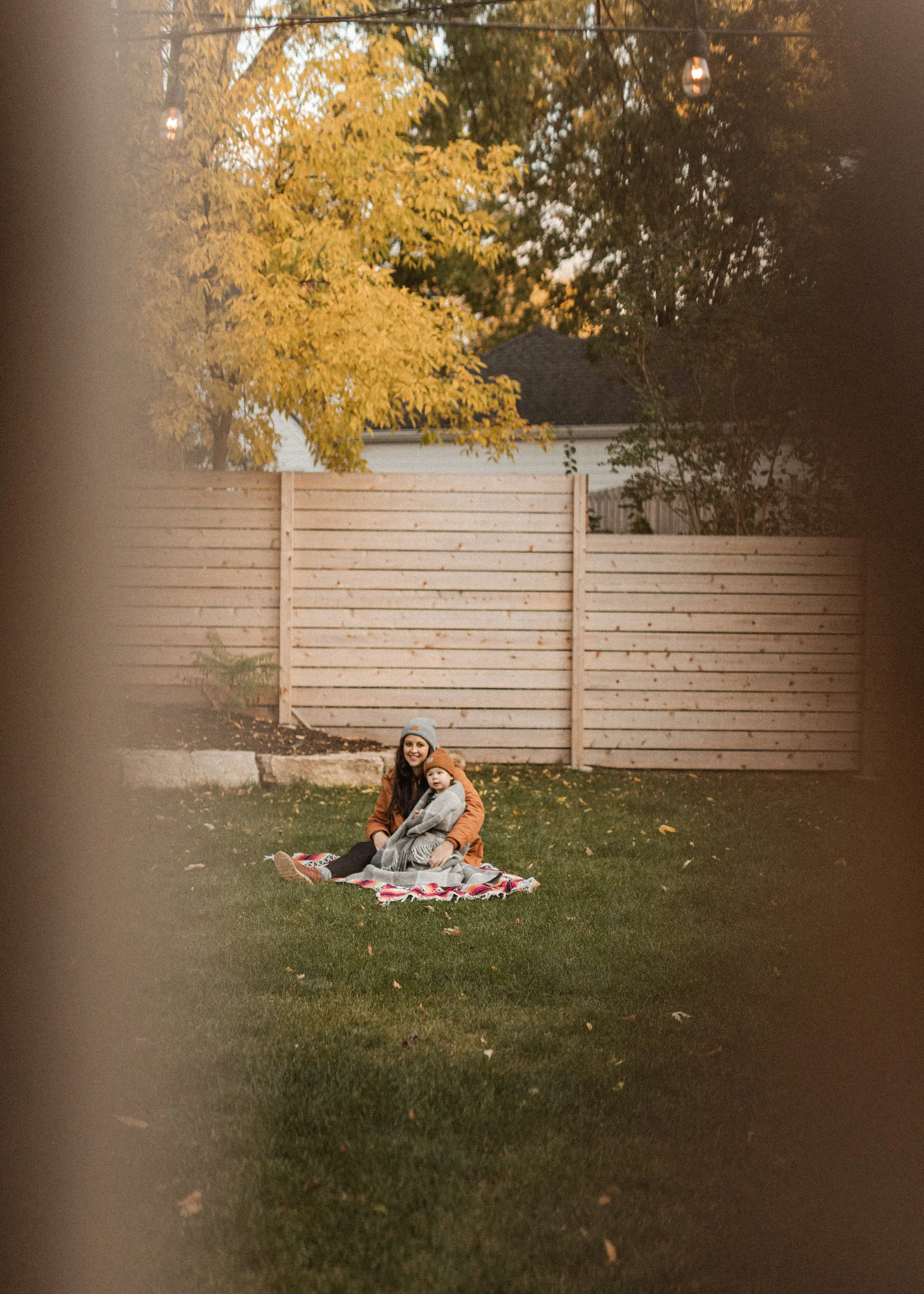 A woman and a child sitting on a blanket in a backyard with a wooden fence and trees with yellow and green leaves, during fall. The woman is smiling and wearing a gray beanie and a brown jacket, while the child is in a gray coat and orange beanie.