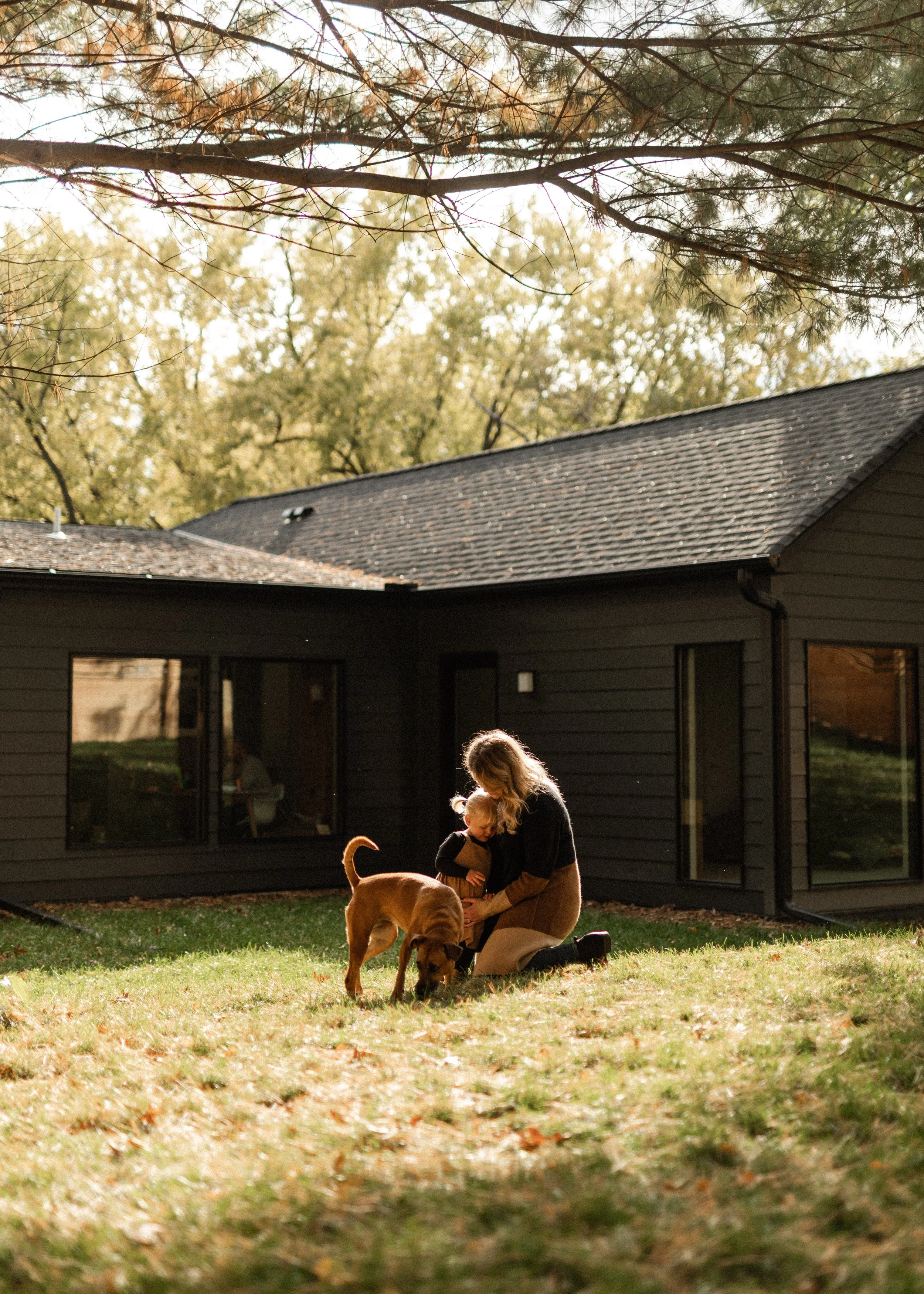 A woman with long blonde hair kneeling on grass with two young children and a dog outside a house with a dark gray exterior, large windows, and a sloped roof. It is fall, with leaves on the ground and trees with yellow and green foliage in the backgr
