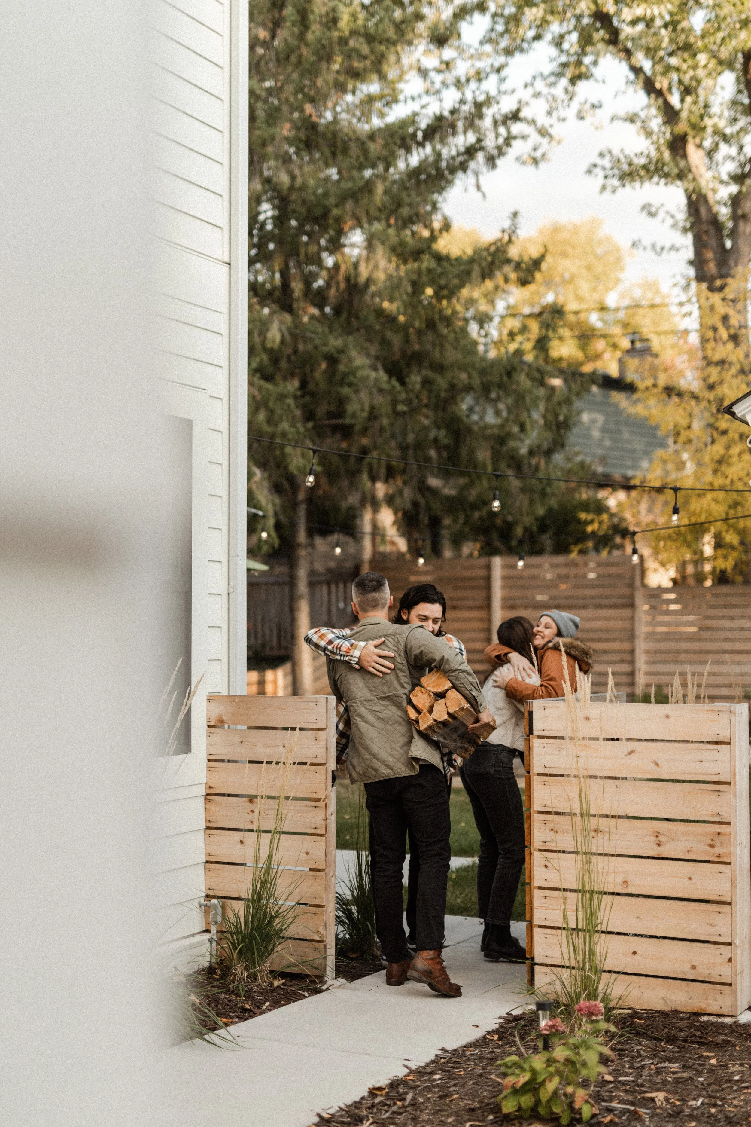 People hugging in a backyard with string lights, wooden planters, and trees with autumn foliage.