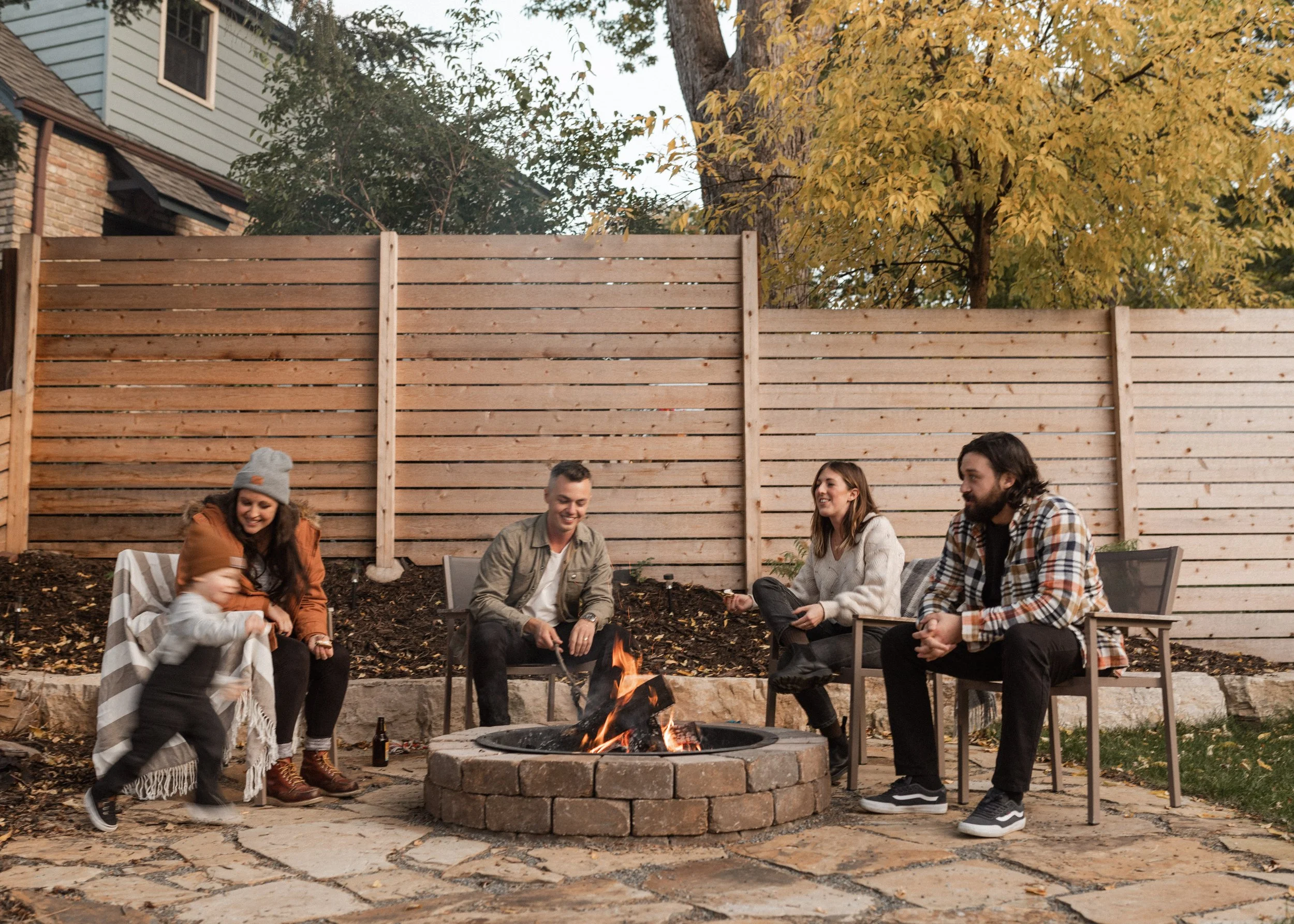 Group of five young adults sitting and standing around a backyard fire pit during autumn, with a tall wooden privacy fence and trees with yellow leaves in the background.