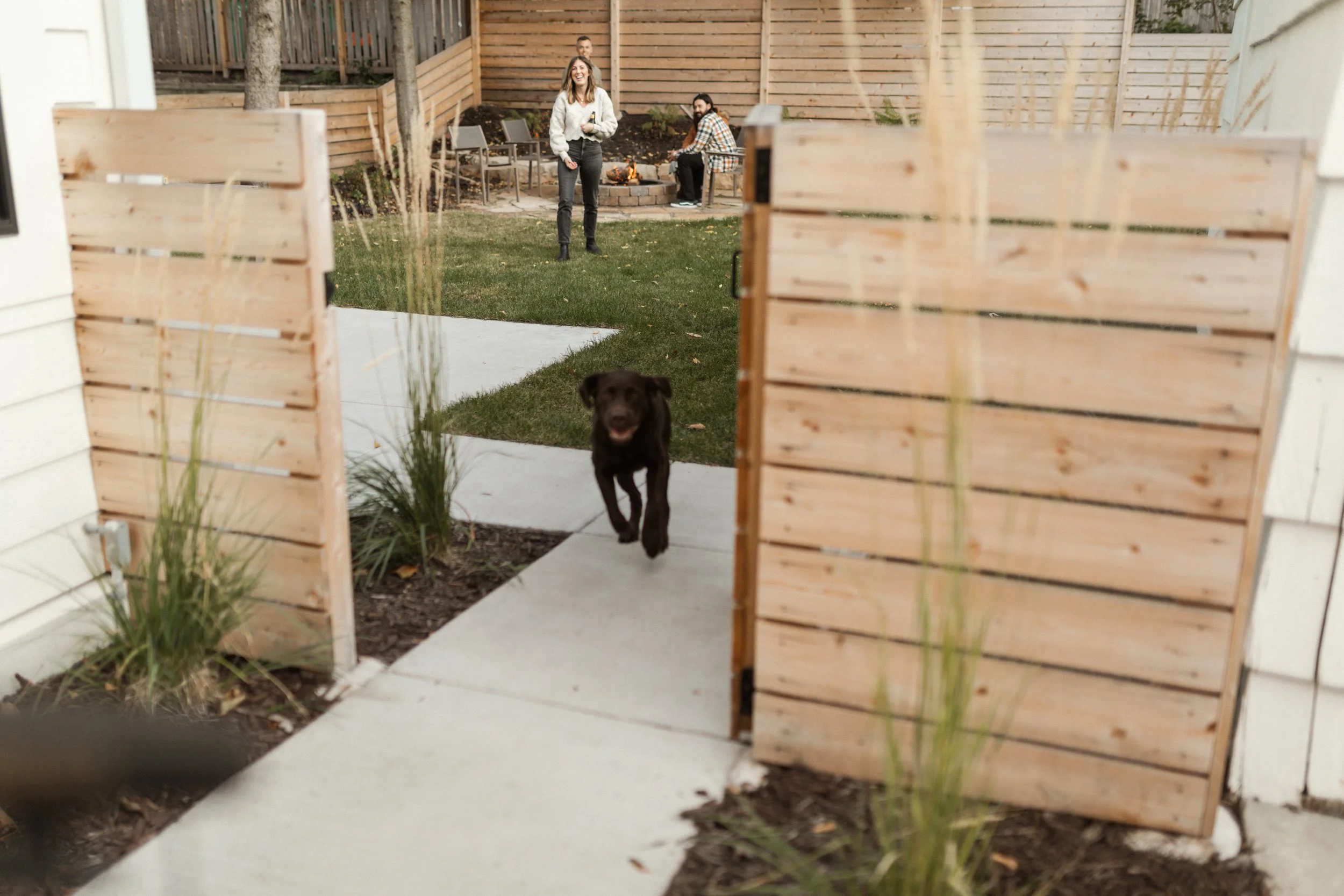 A person smiling, standing on a lawn, with a dog running towards the camera, seen through an opening in a wooden fence. Two other individuals are sitting near a fire pit in the background.