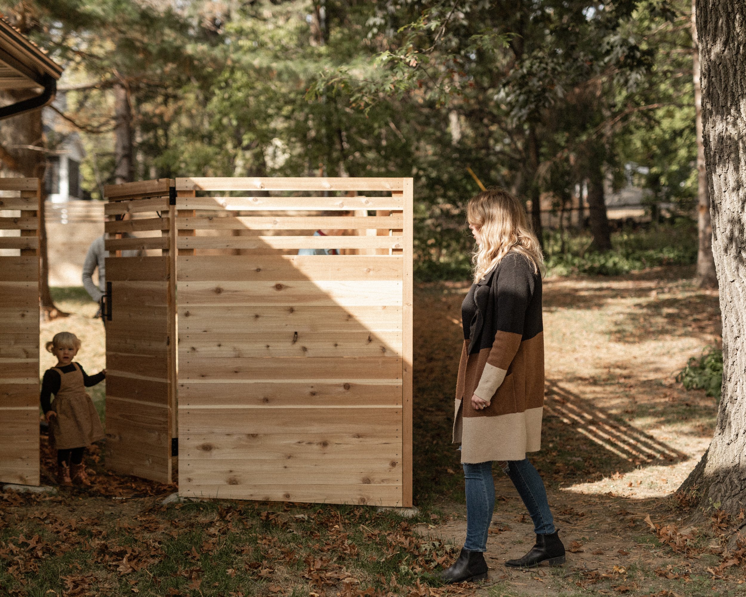 A woman with blonde hair, wearing a black jacket and brown skirt, stands outdoors near a tree, looking at a wooden structure made of light-colored wood slats, with a child nearby in a brown dress and black top, and another person partially visible be