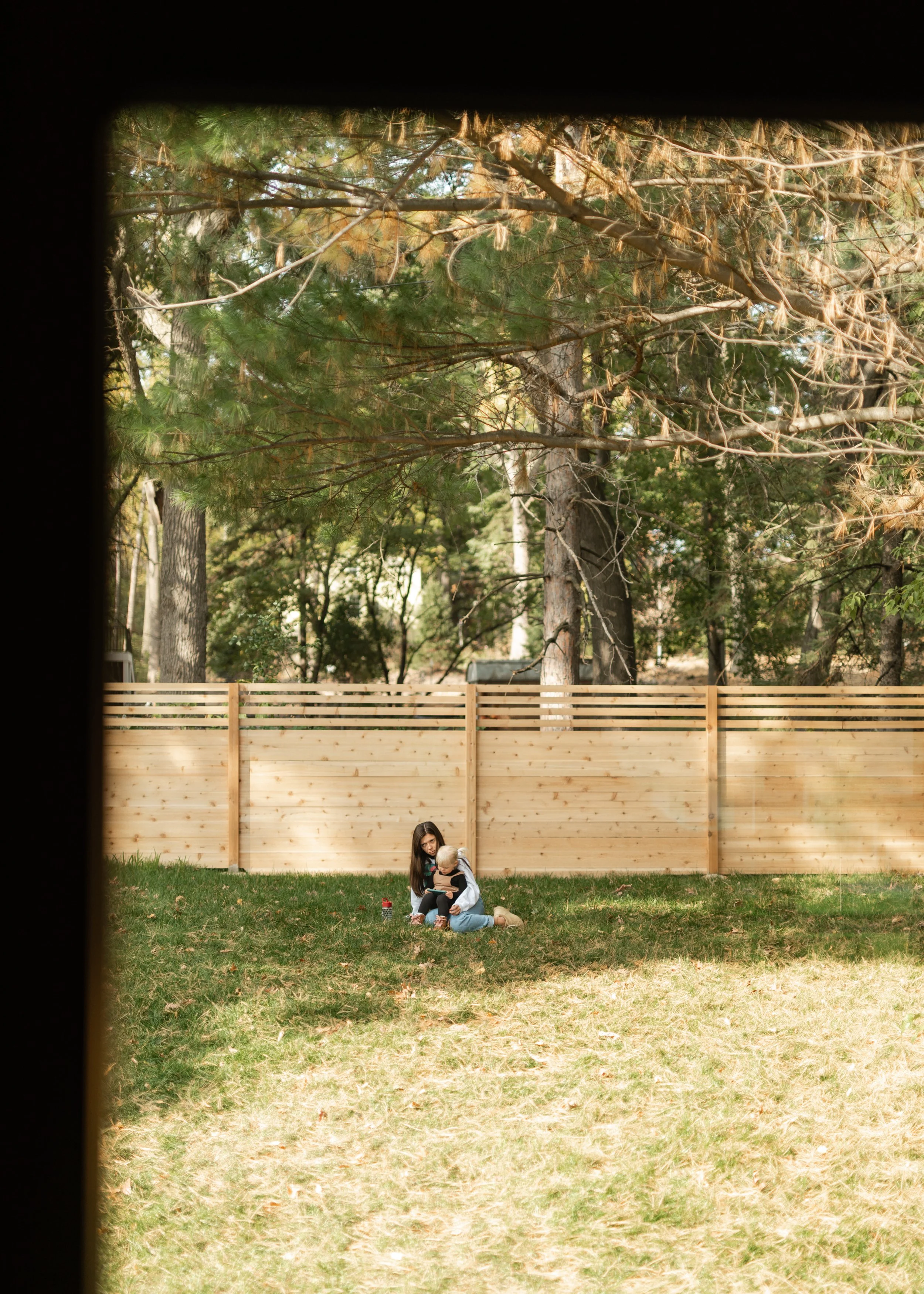 A woman and a young child sitting on the grass in a backyard, with a wooden fence and tall trees in the background.