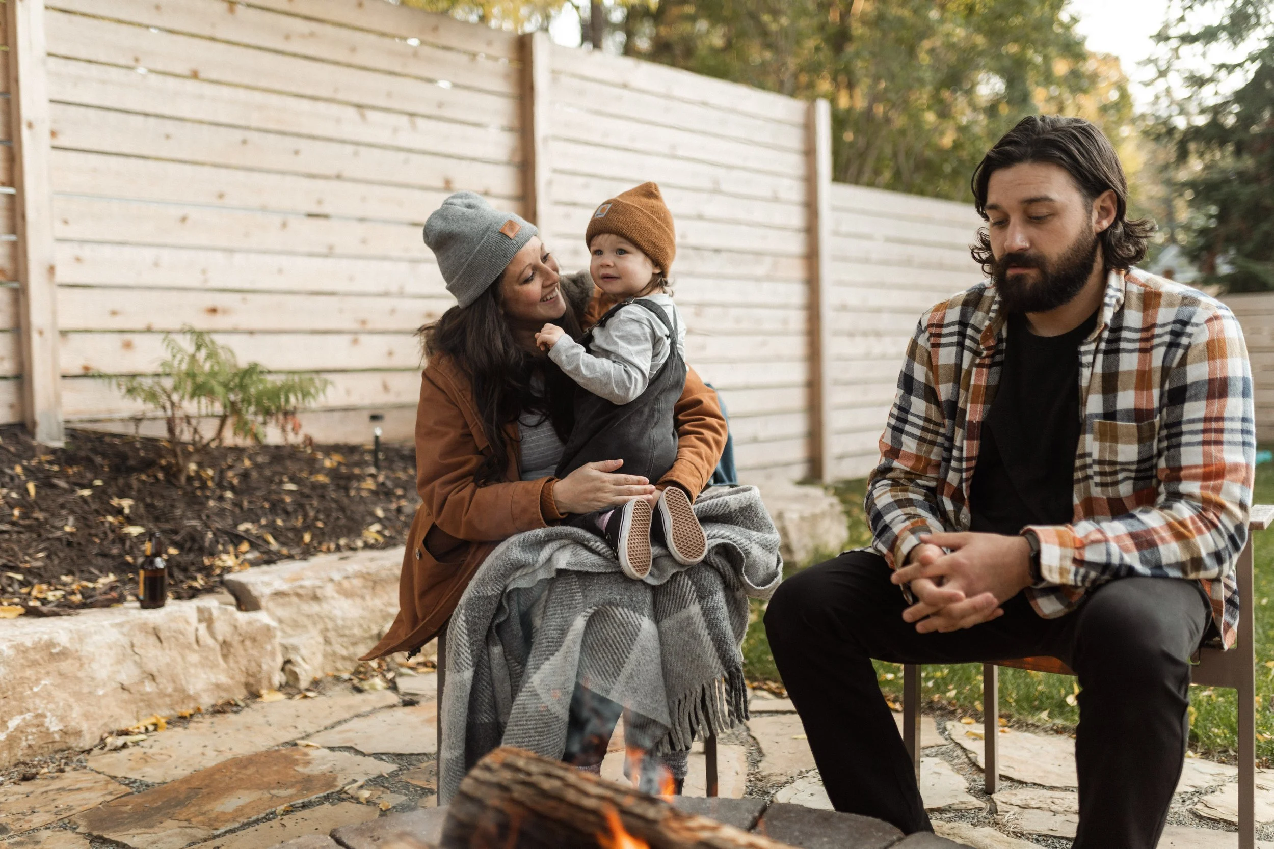 Family sitting outdoors near a fire pit, enjoying a fall day. The woman is holding a young child, and a man sits nearby. They are dressed in warm clothing, with trees and a wooden fence in the background.