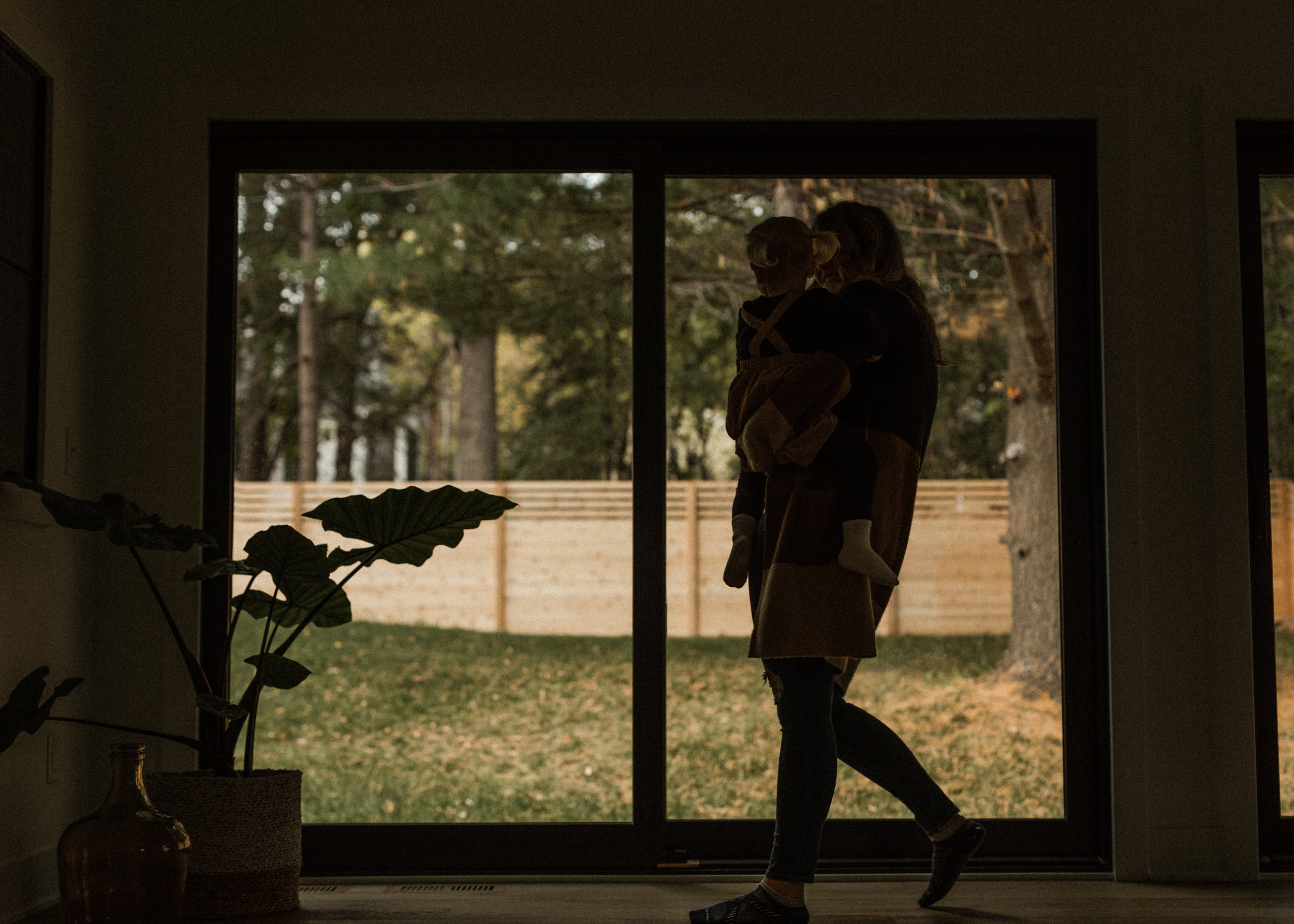 A silhouetted woman is holding a small child in front of a large window looking out onto a backyard with trees and a wooden fence. The scene appears to be during the evening or late afternoon.